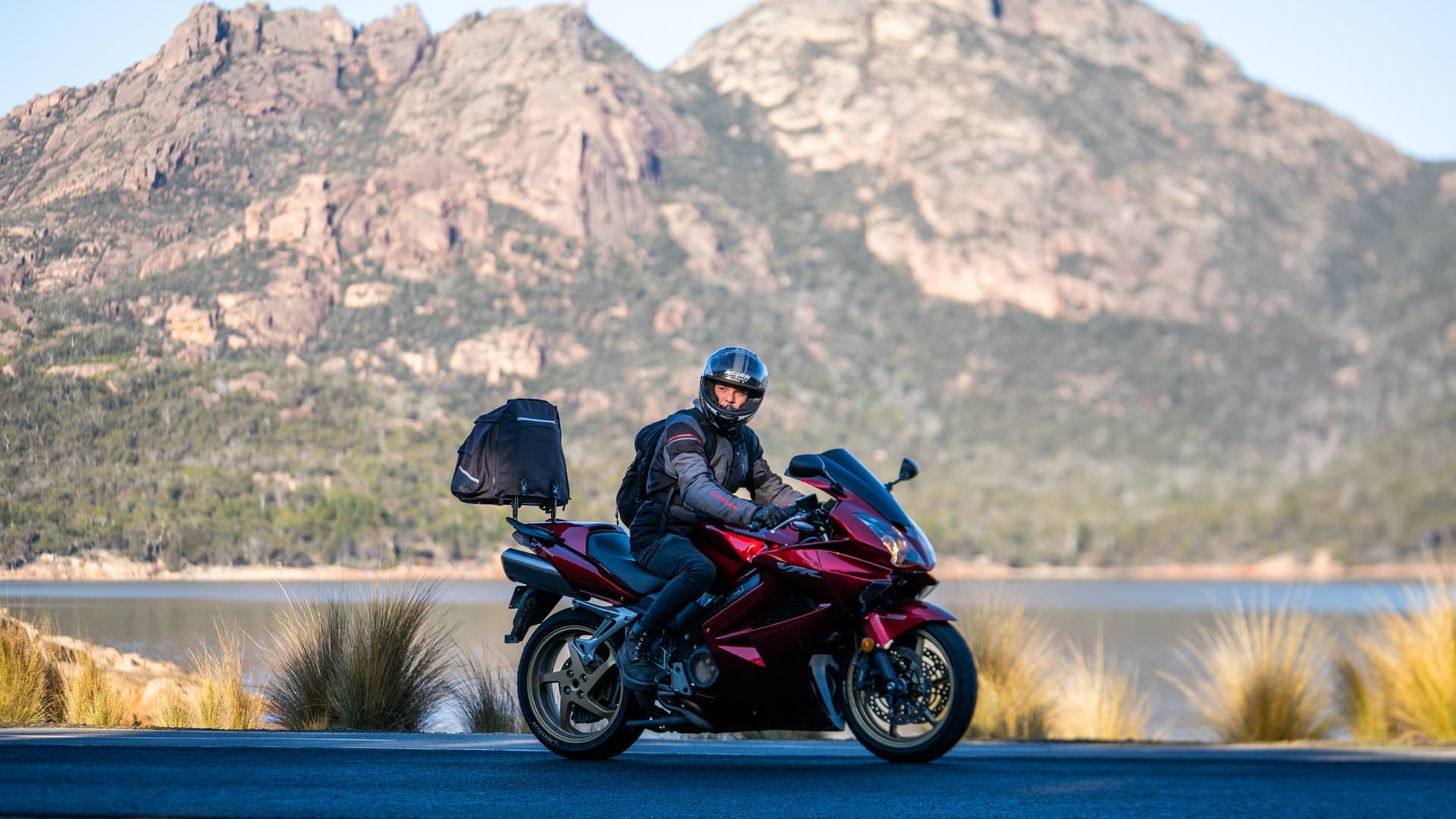 Declan riding a red motorbike beside a lake and mountains in Tasmania