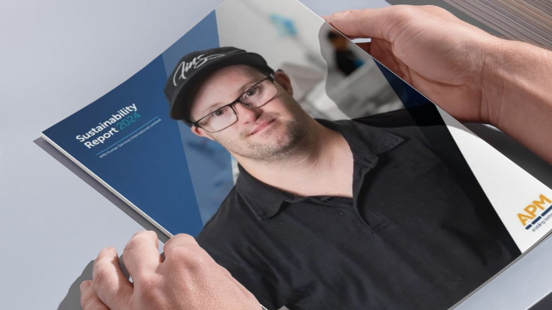 APM Sustainability Report as a printed document with hands looking through. The cover has a young man with Down syndrome