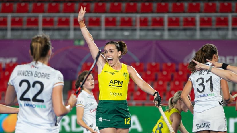 Australian women’s hockey team player in yellow and green uniform raising an arm in celebration during a match against opponents in white uniforms. APM and Ingeus logos are visible on the uniform. Red stadium seats and purple banners in the background.