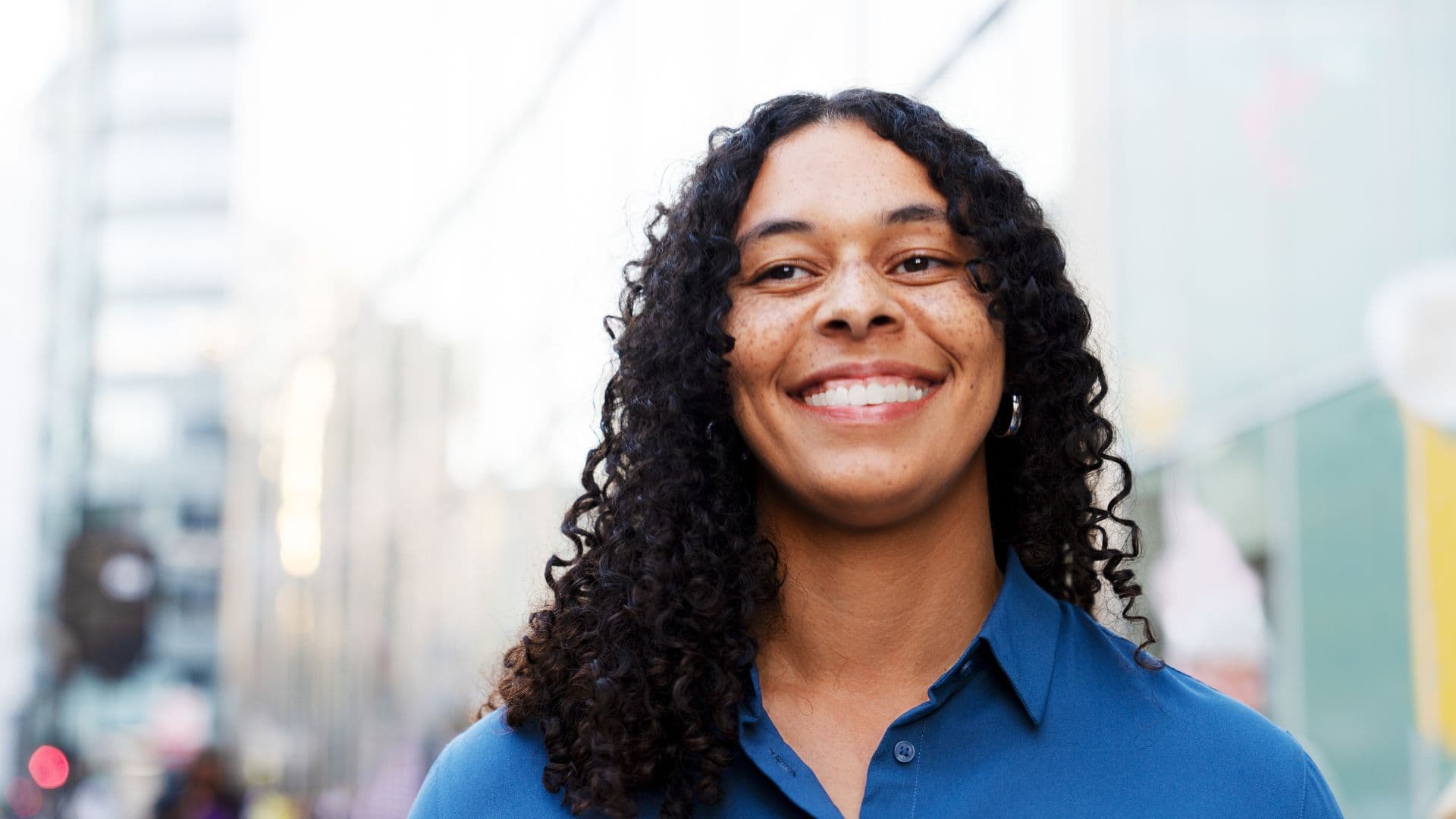 Woman smiles in street with dark curly hair and blue top
