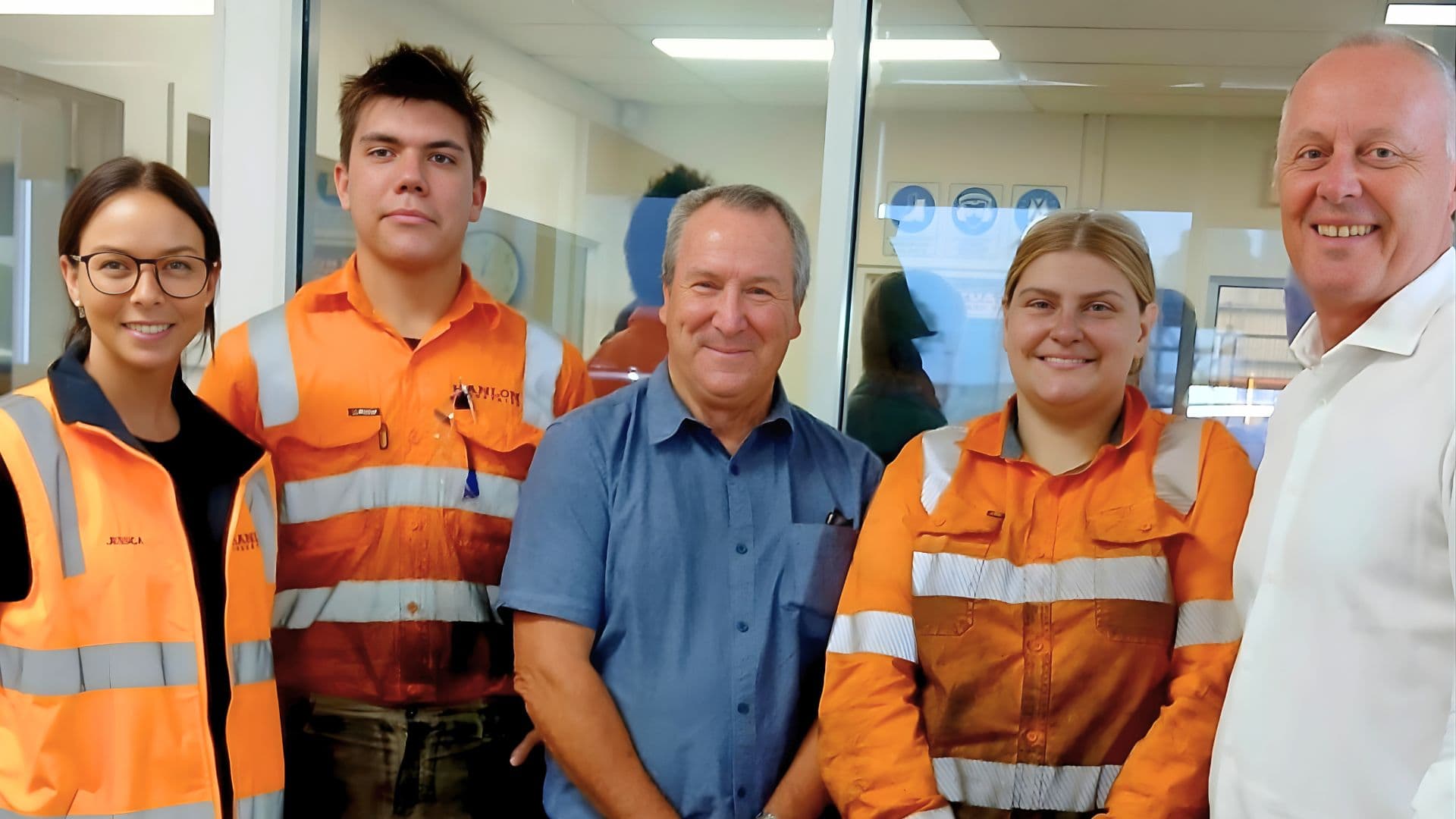 Australian Apprentices Mahlie and Vernon wearing high-visibility orange work uniforms, standing with colleagues in a workplace setting.