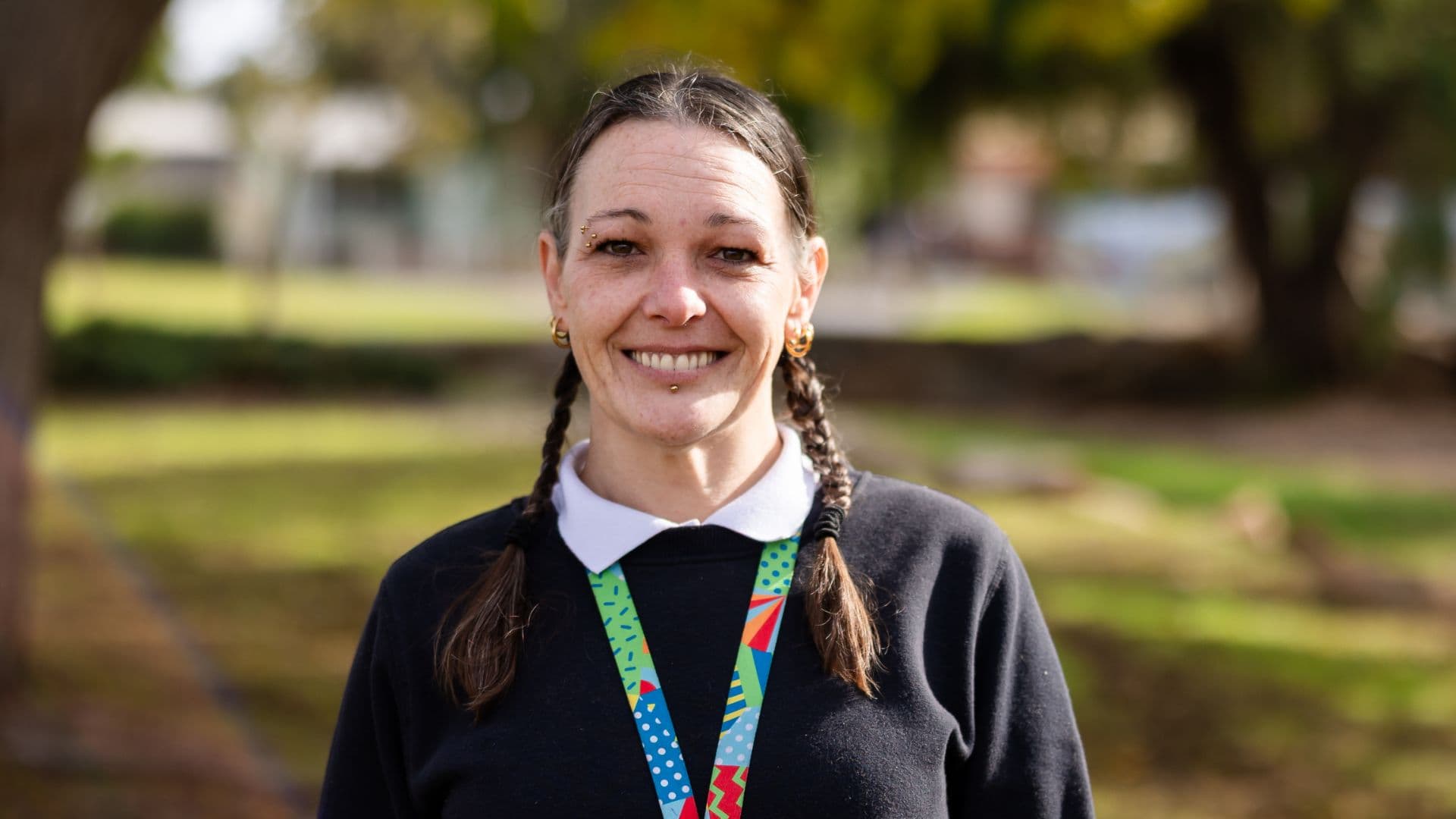 Kylie stands outside with greenery in the background, wearing a dark sweater and colourful lanyard, representing her success story with APM Workforce Australia.