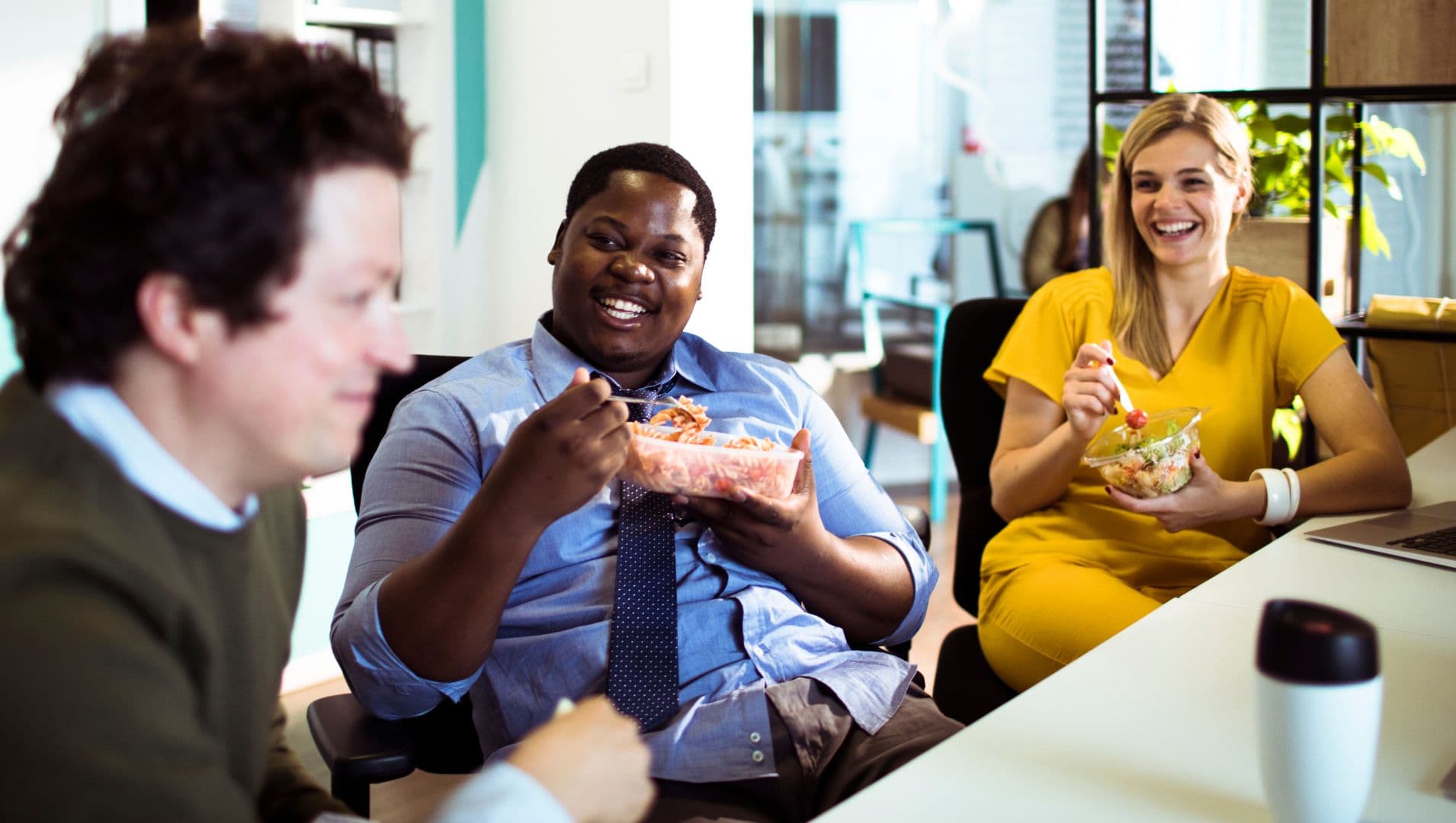 Three colleagues smile while eating lunch at their desk