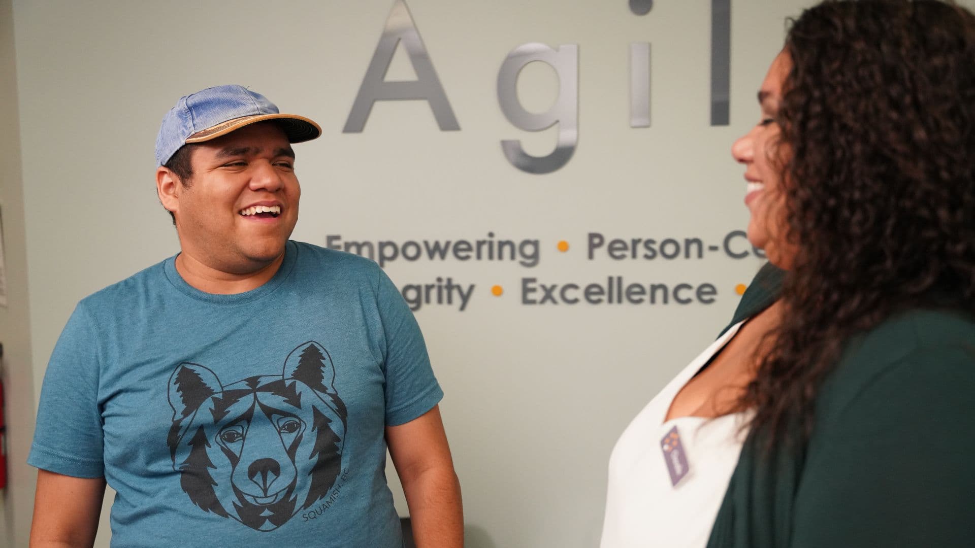 Two people standing in an office environment, smiling and conversing near a wall with the text " Empowering - Integrity - Excellence." One person is wearing a blue t-shirt with a bear design and a cap, and the other is in a white blouse.