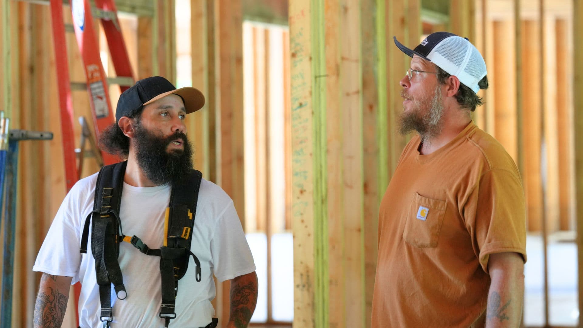 Two construction workers in conversation on a building site, surrounded by wooden beams and construction material. One worker wears a harness and a cap, while the other sports glasses and a cap
