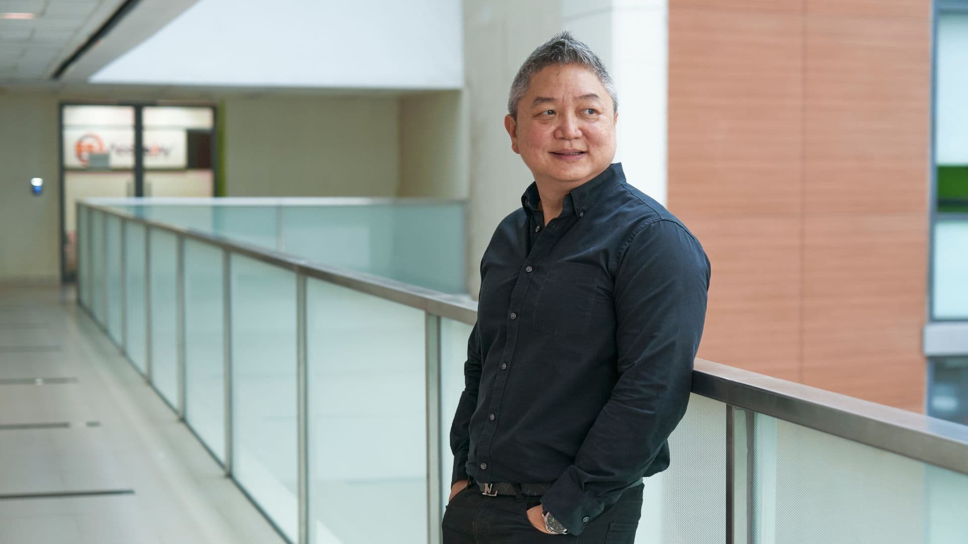 Jek standing in a bright office corridor, hands in pockets, wearing a black shirt. This image represents his journey with Ingeus Singapore, where career coaching and resume refinement helped him return to the workforce.