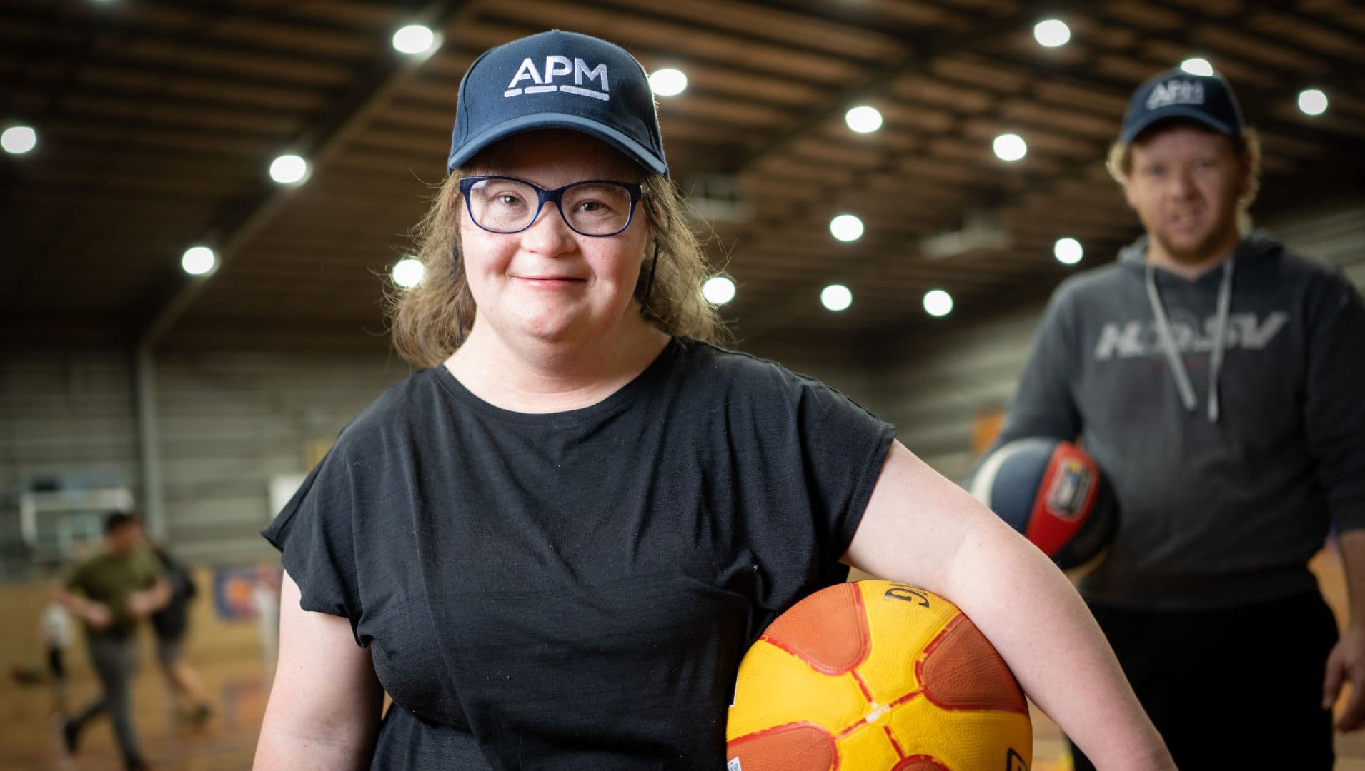Holly holds a basketball wearing an APM cap inside a community gym