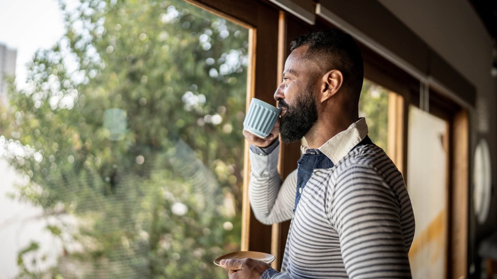 A man drinking a cup of coffee in his home and looking out a window