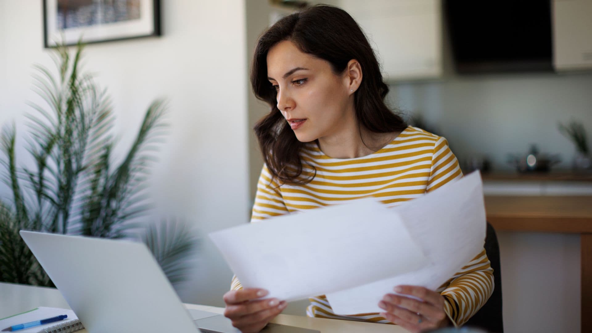 A woman sits at a table reviewing paperwork while looking at a laptop, holding documents in her hands in a quiet home workspace.