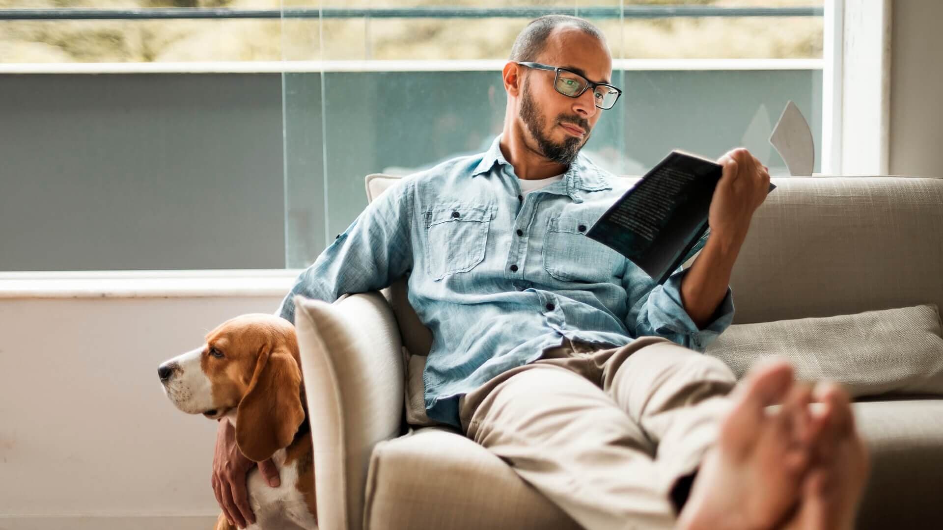 A man reading a book on the couch with his dog next to him