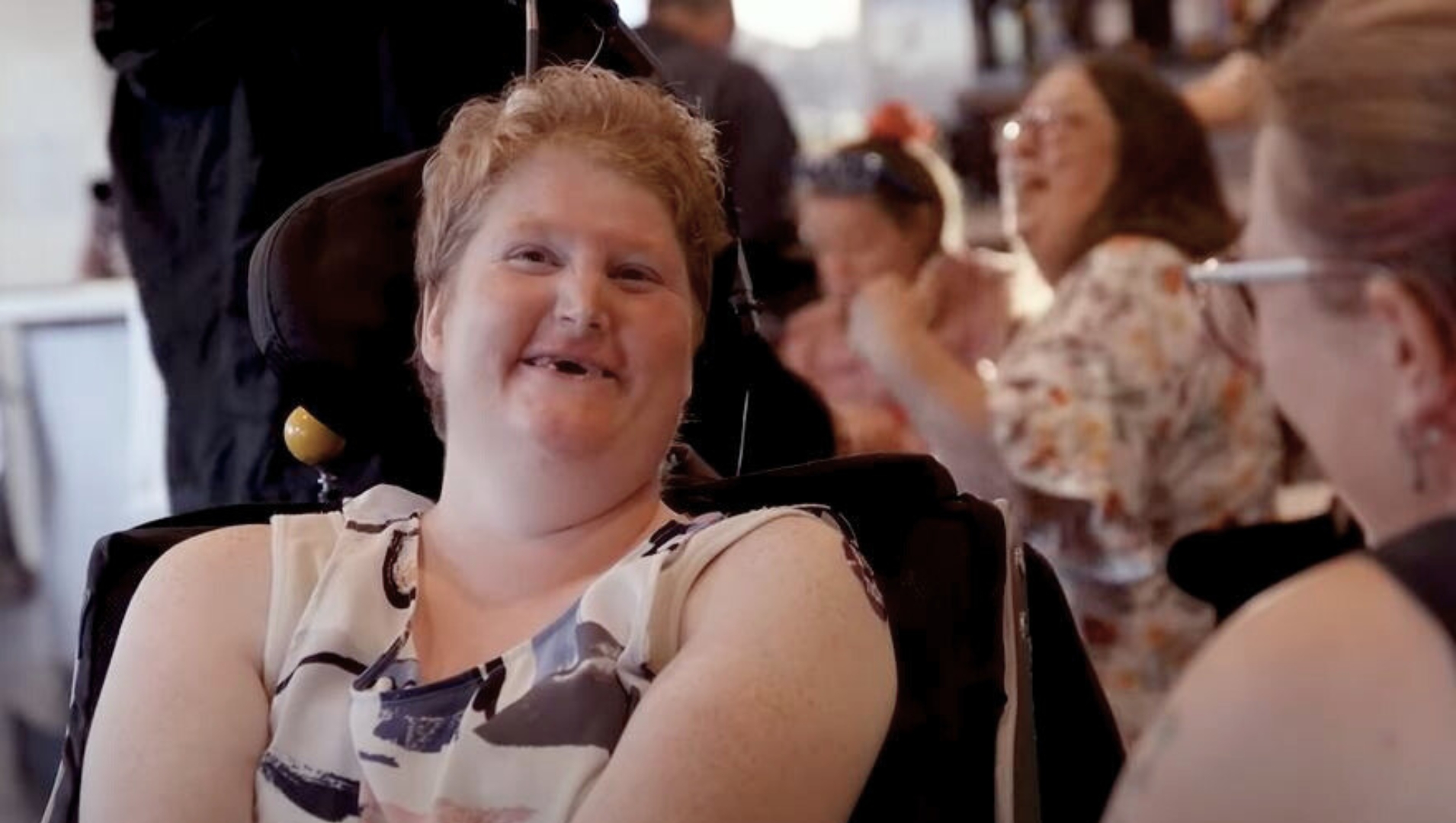 A woman in a wheelchair smiles with a friend at APM Communities EasyBeatz disco for people with disability