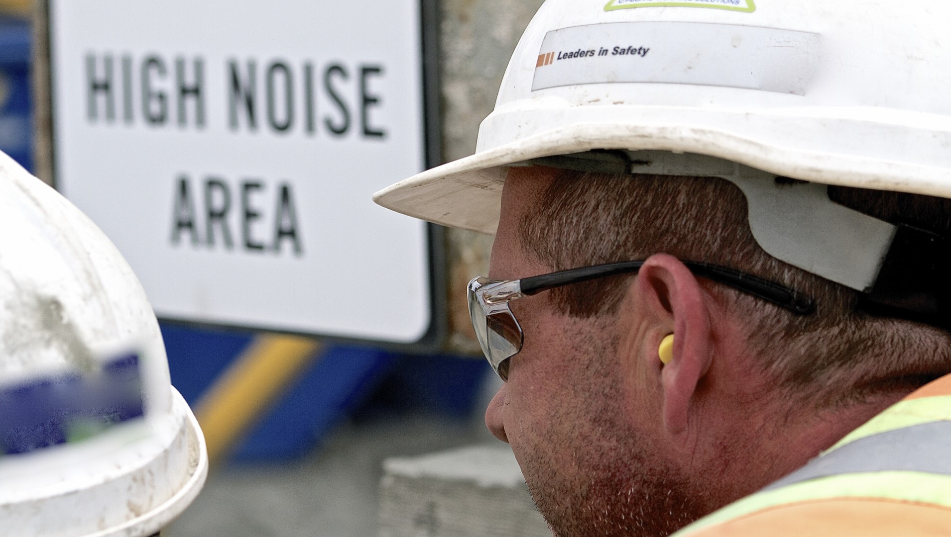 Hearing impaired construction worker at a job site, with earplugs in, in front of a sign which reads "High Noise Area"