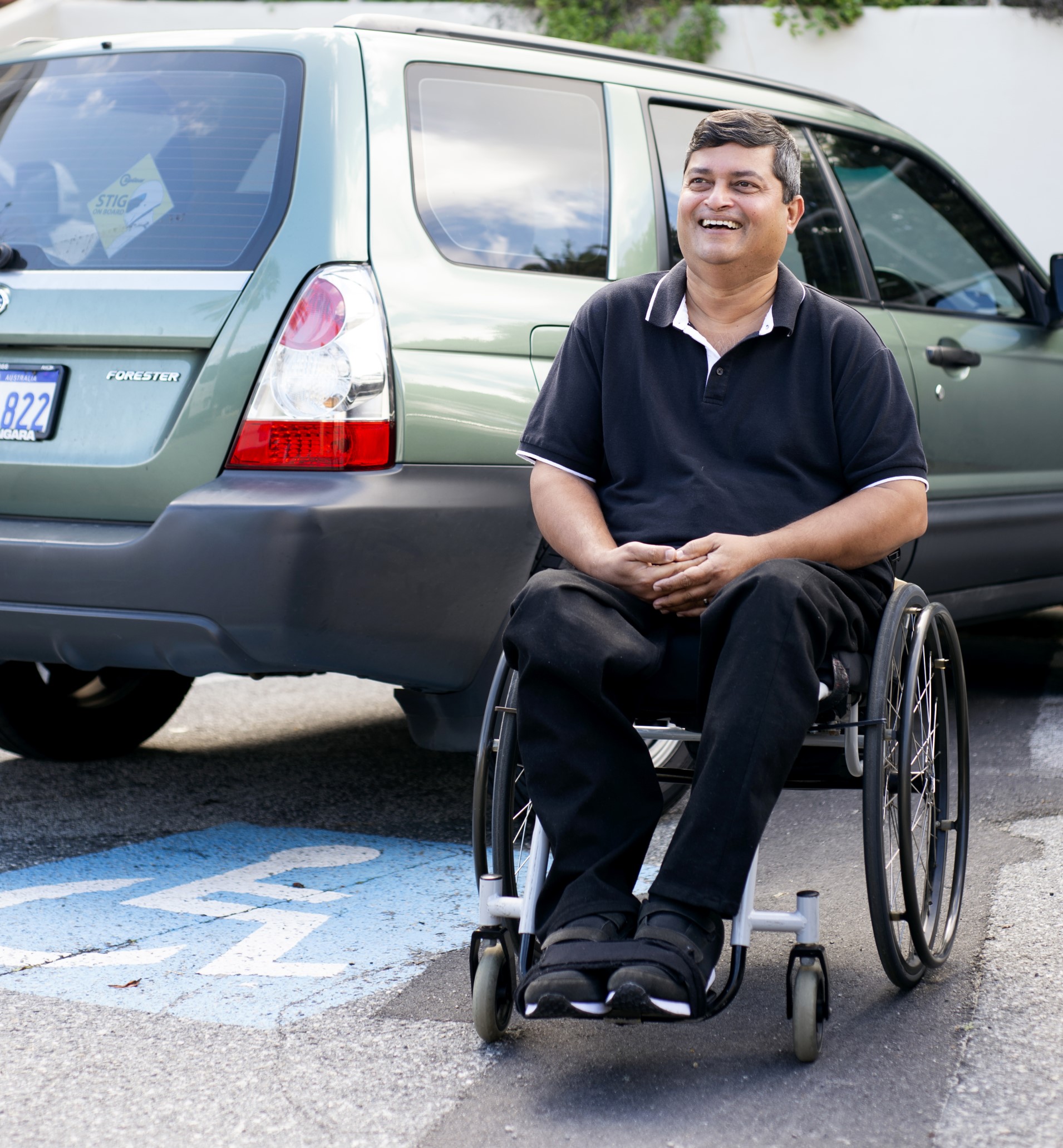 A man with dark hair, wearing all black, sitting in his wheelchair in front of a car