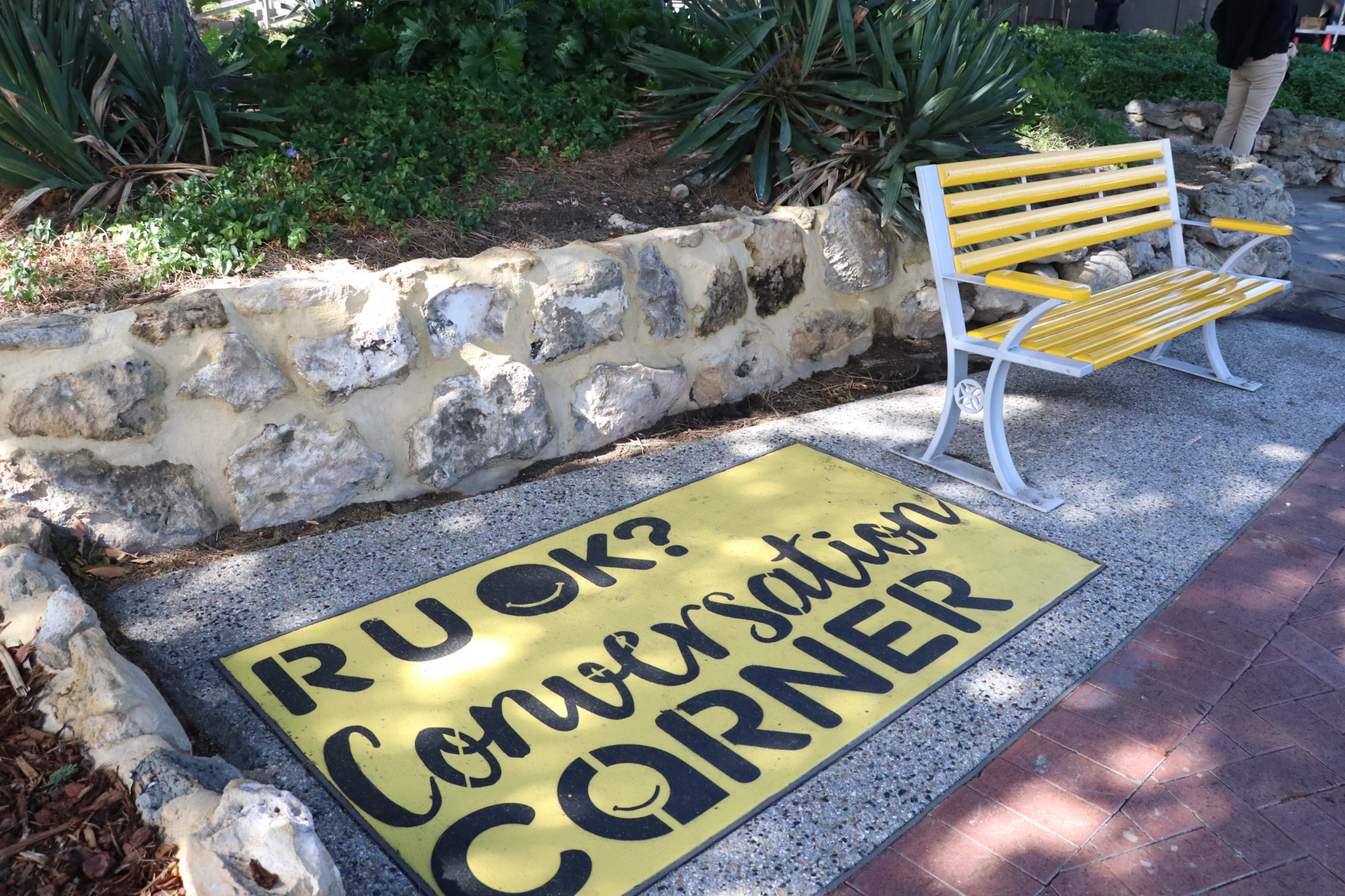 A yellow bench next to a sign painted on the ground that reads: R U OK? Conversation corner