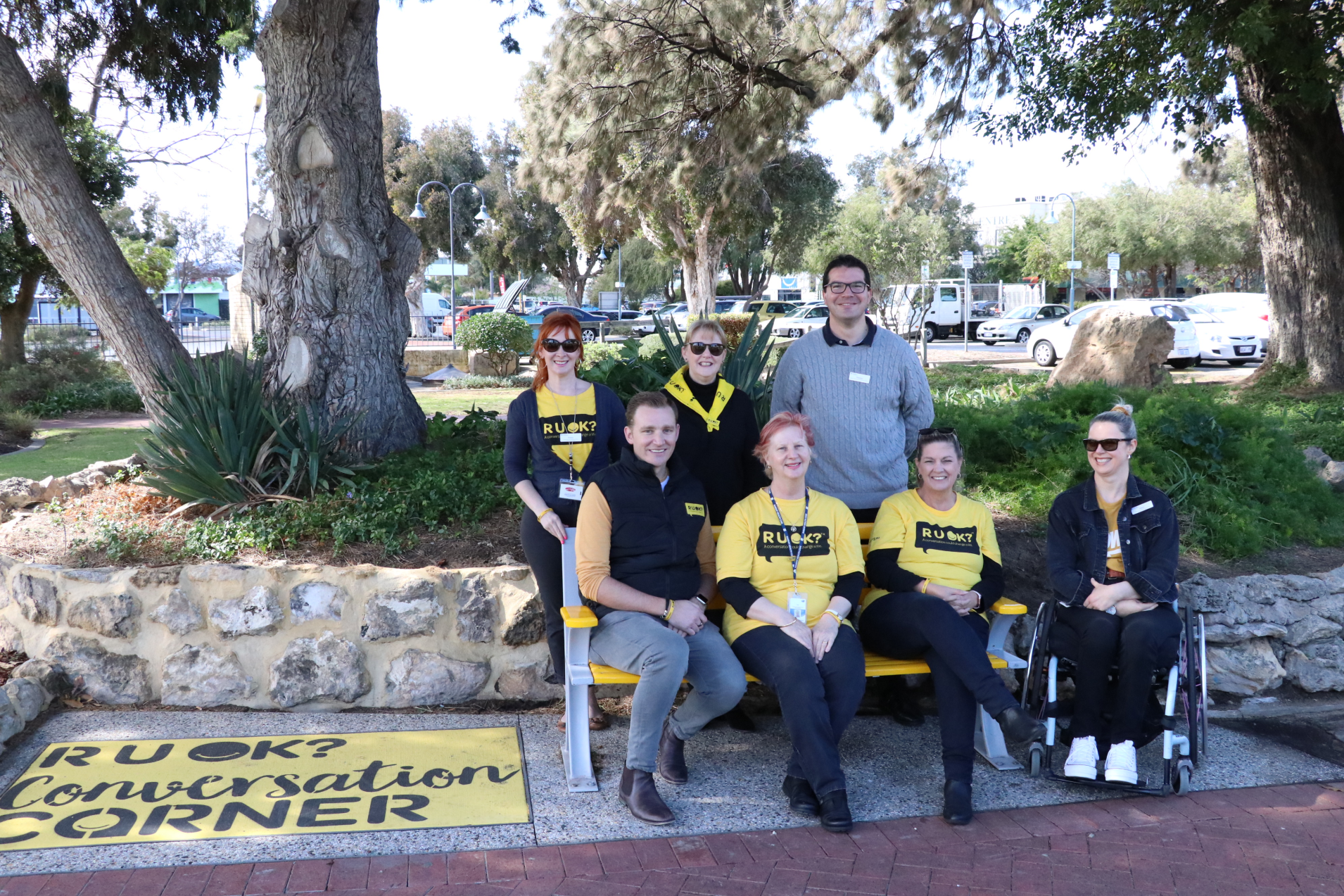 A group of people seated on and around the yellow bench in the park