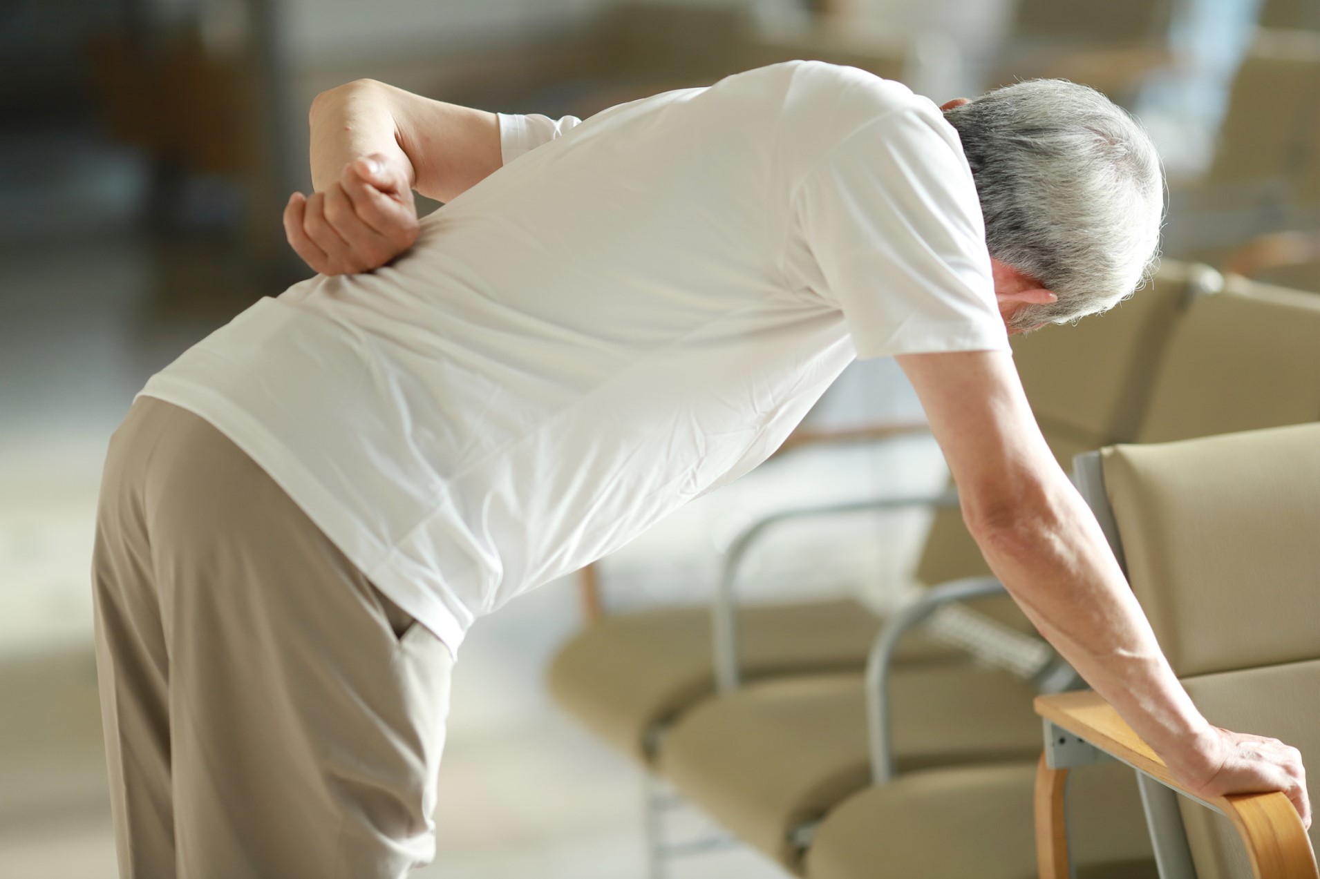 an older man with spinal condition, bending over, clutching his lower back, grimacing in pain