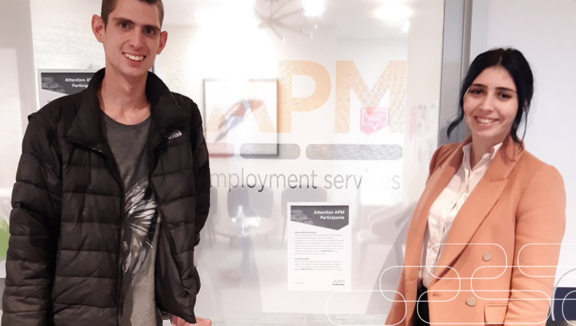 Aaron standing with his APM Employment Consultant April in front of a door with the APM logo on it