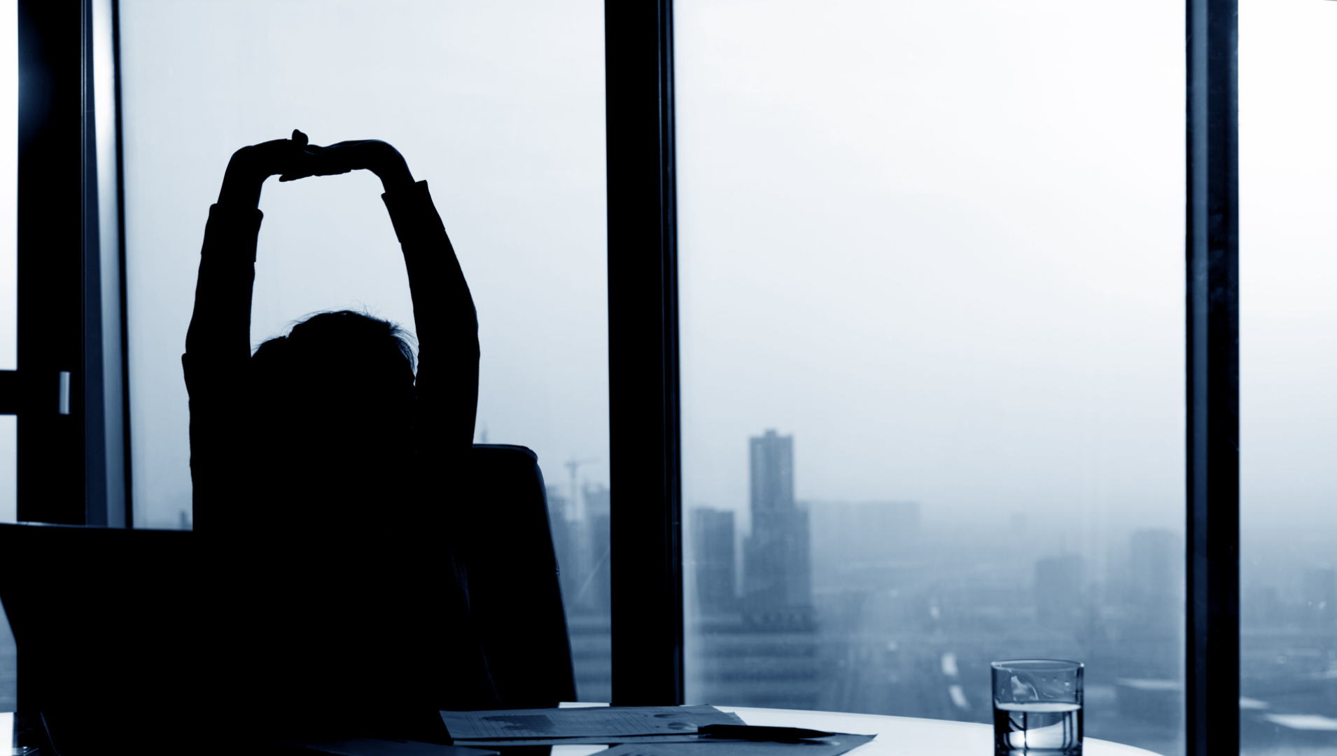 Silhouette of a woman stretch her arms above her head at her desk in front of a window