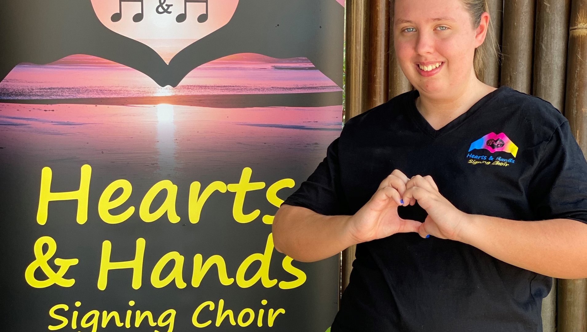 Xanthe standing beside a 'Hearts and Hands' banner, holding her hands in the shape of a heart