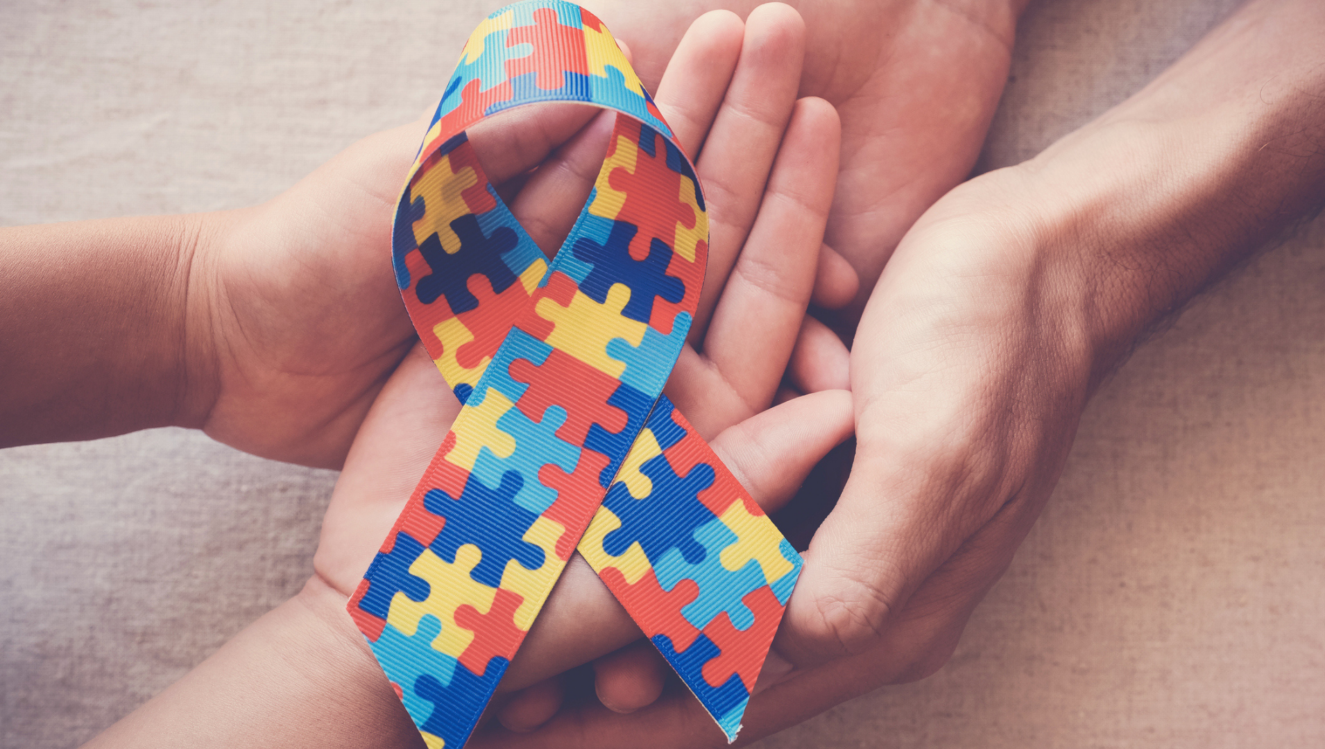 A pair of hands framing the autism awareness ribbon, coloured with orange, dark blue and teal puzzle pieces