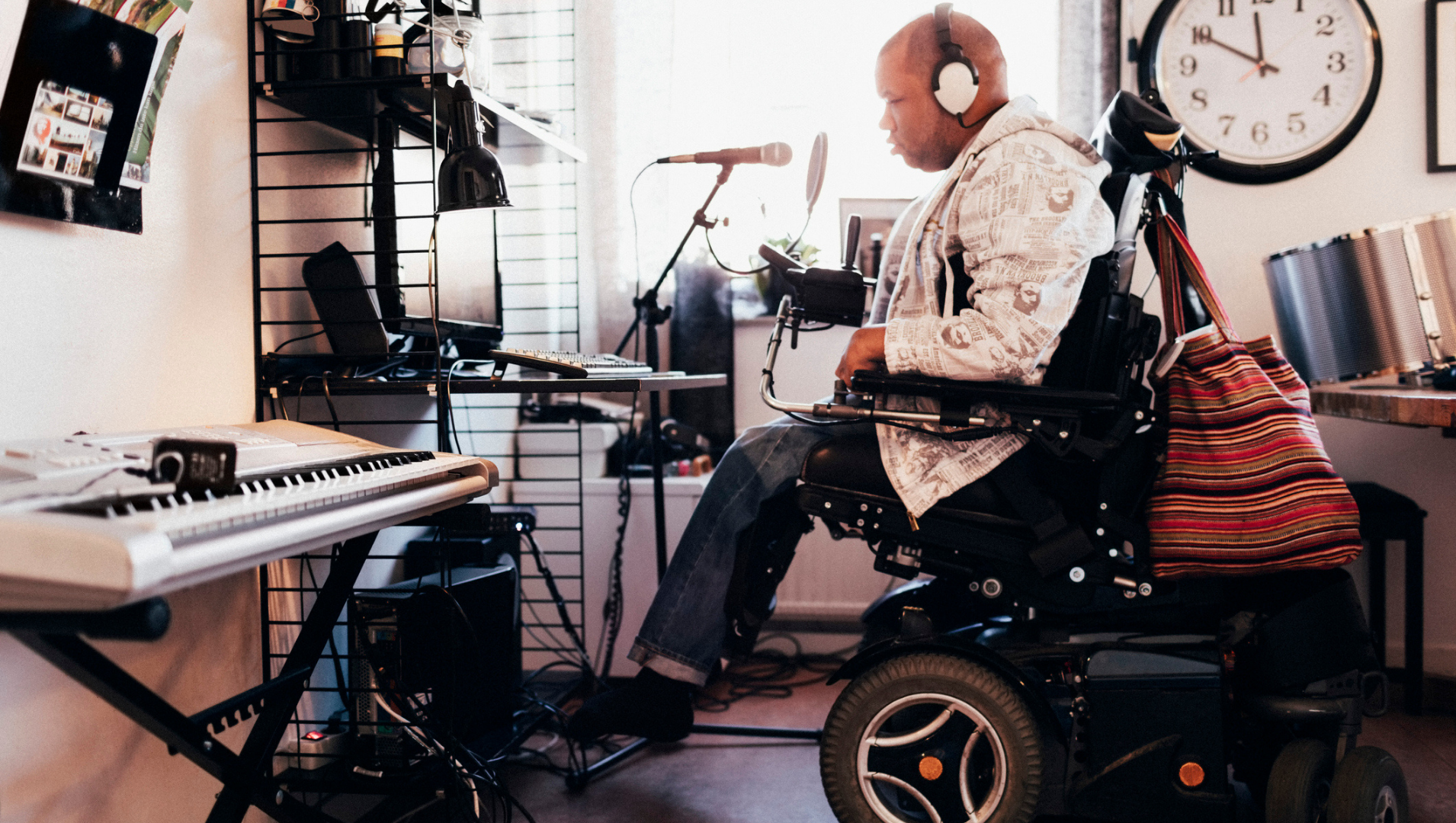 A man in a motorised wheelchair is sitting in a music studio with headphones on, in front of a microphone and a keyboard