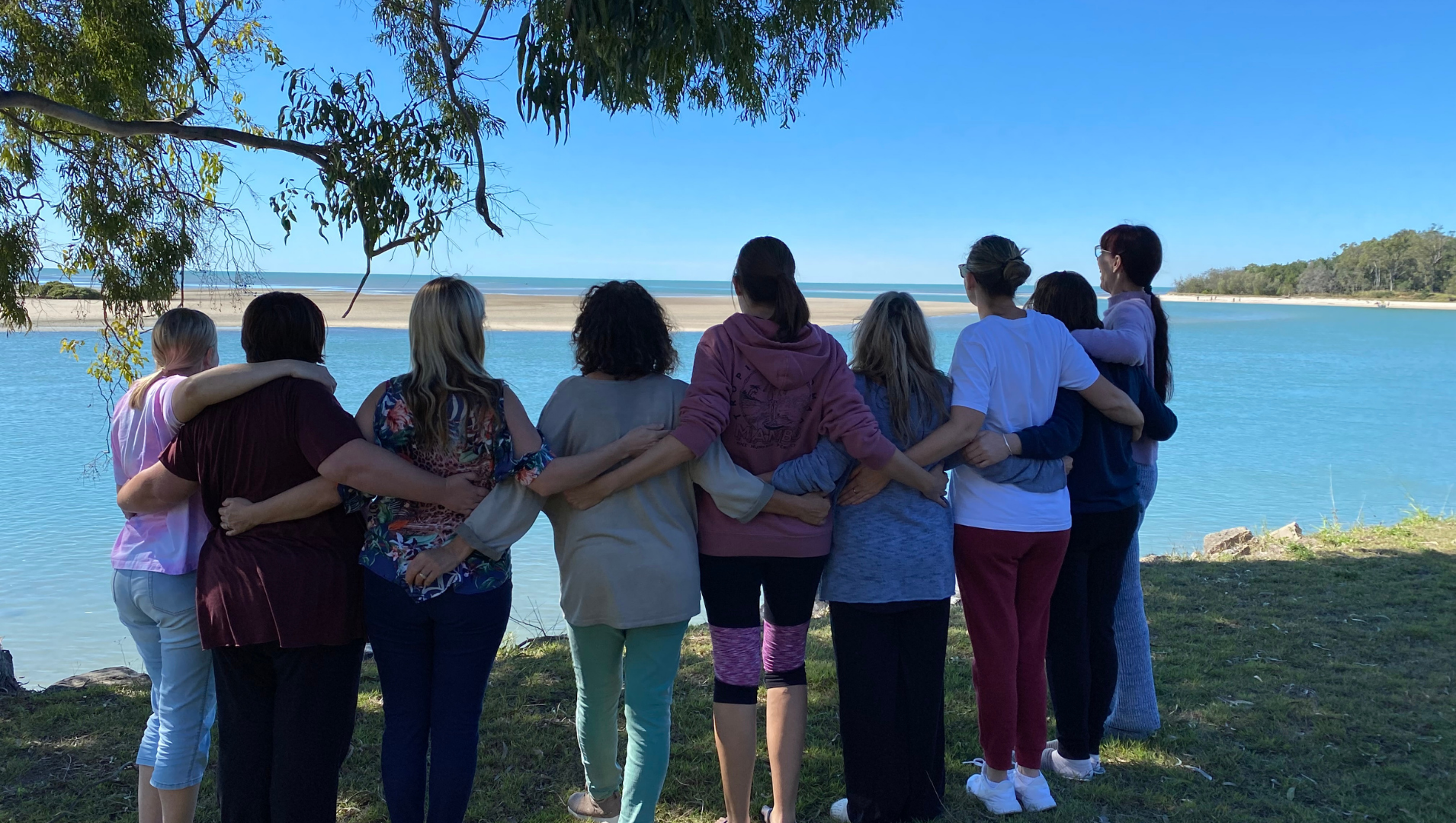 It's a bright sunny, clear day and 9 women stand facing a glistening body of water, embracing by wrapping arms around each others' waists