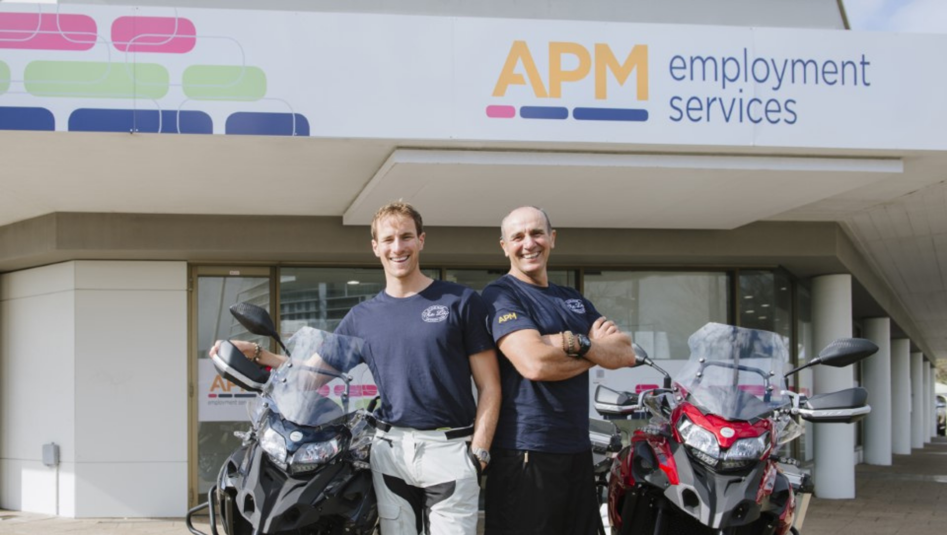 Joel and his dad standing back to back smiling in front of an APM office