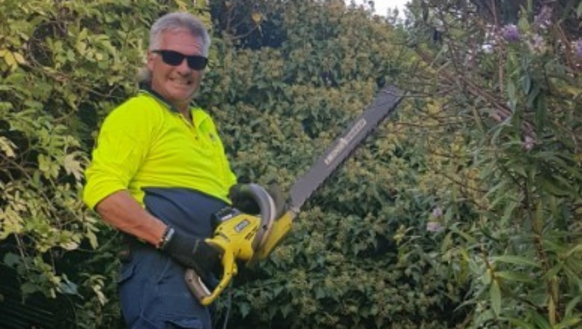 Wayne is pictures tanding in front of an overgrown tree, in his hi-vis work uniform and PPE, holding a chainsaw and smiling