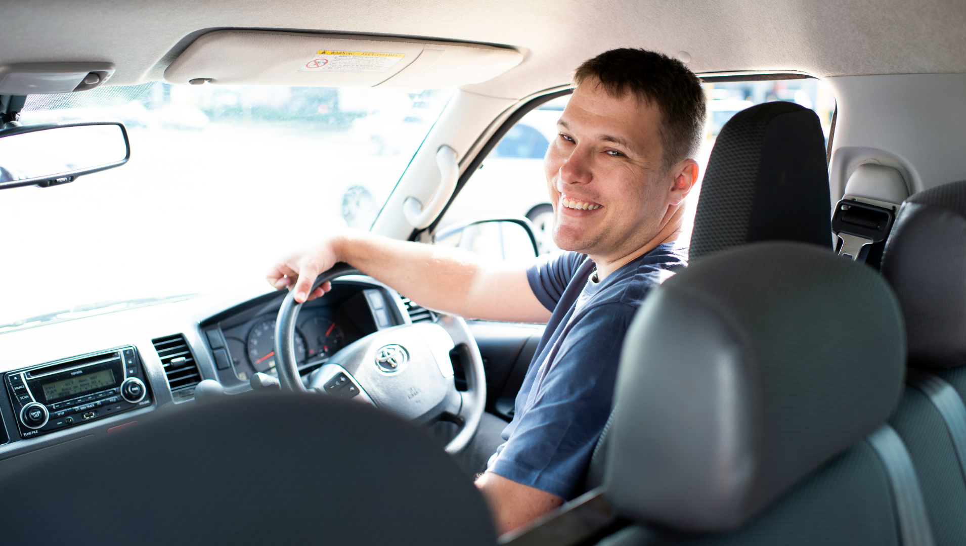 Alex pictured sitting in the front seat of a work can, in the driver's seats, smiling to a person in the back