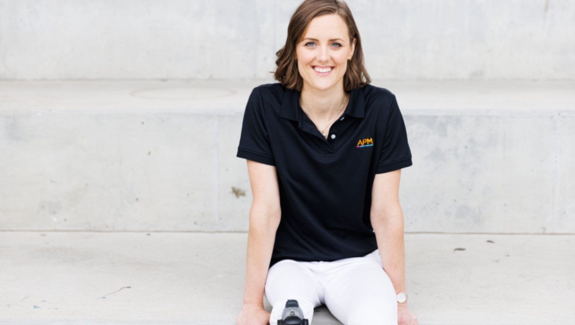 Ellie Cole, wearing an APM poloshirt is sitting on a limestone step, smiling