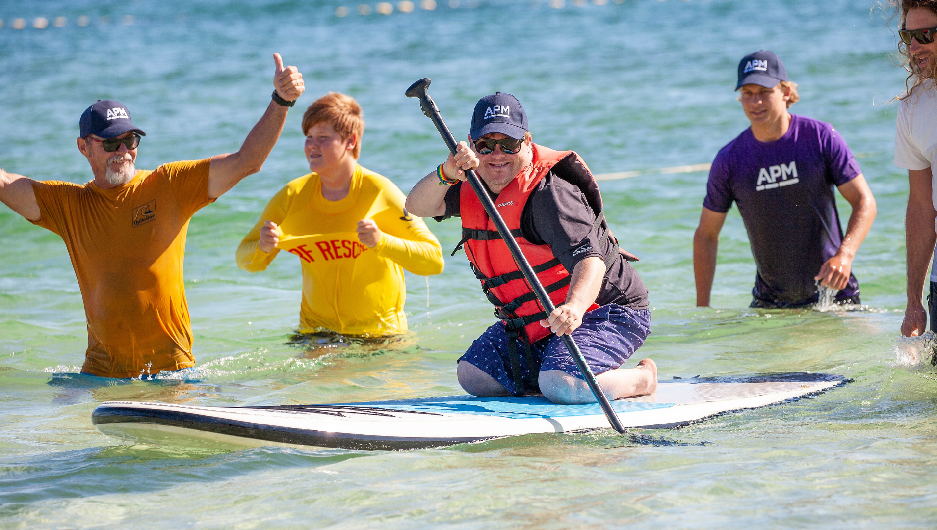 Jack kneeling on a SUP board, floating on the water, surrounded by three team members