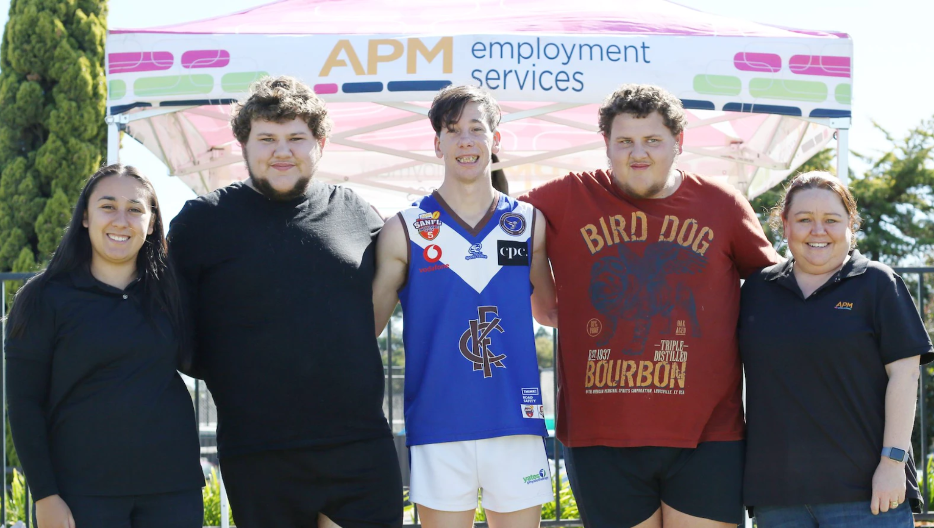 Two APM employees and three SANFL players pose smiling in front of an APM branded marquee