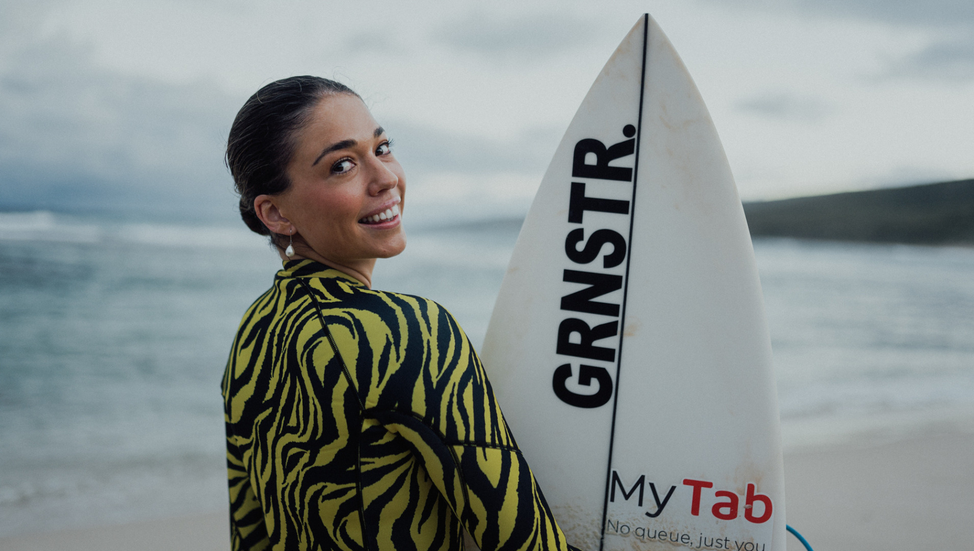 Eliza is pictured wearing a brightly patterned yellow and black wetsuit in front of the ocean
