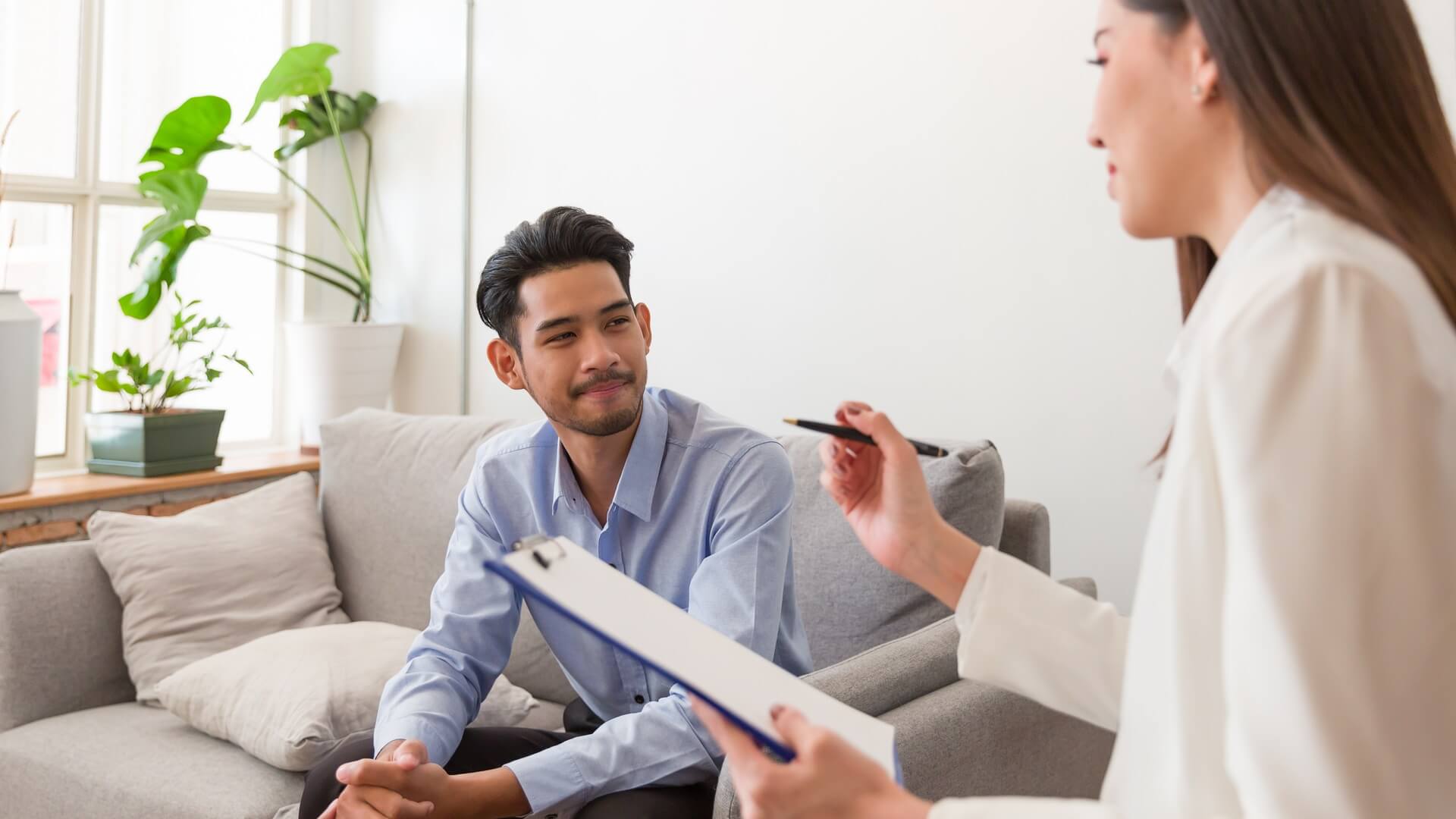 Young man in a blue business shirt sits on a grey sofa with open body language, listening to a female doctor speaking while holding a clipboard.