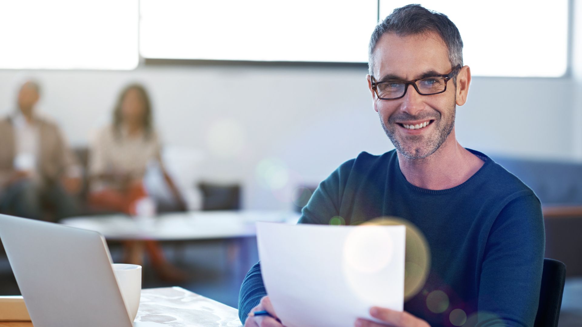 A man smiles at a desk doing admin work on a laptop