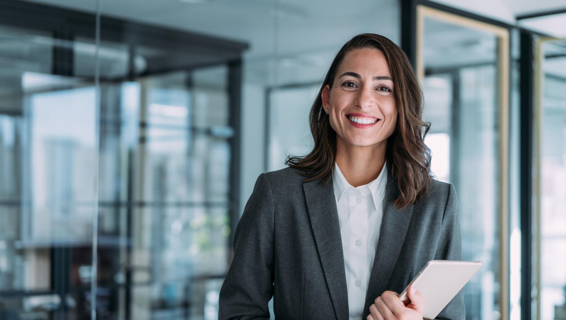 A woman in a suit smiles holding a folder in a bright modern office