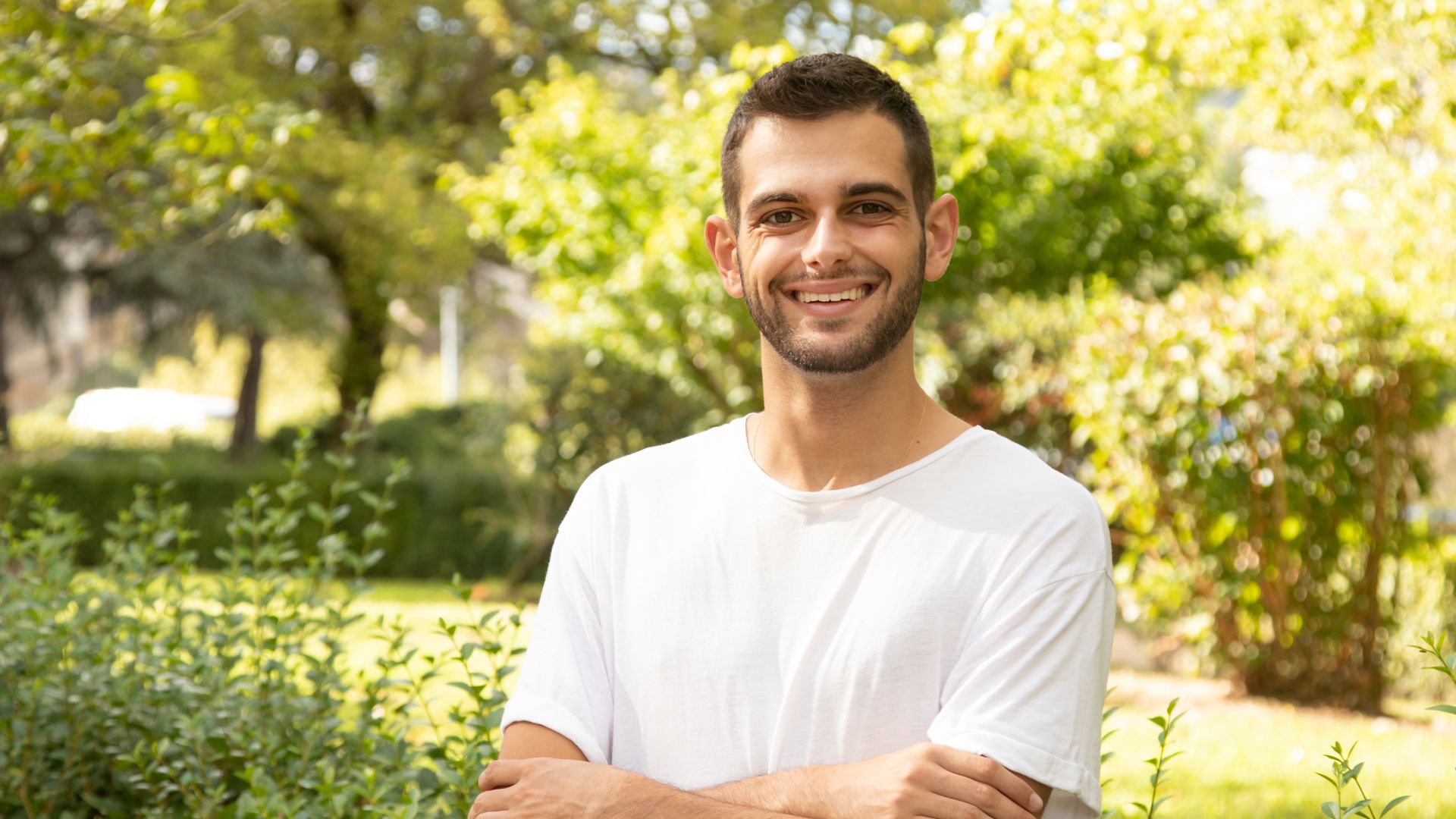 A man with a beard wearing a white t-shirt sitting outdoors, smiling at the camera with trees in the background.
