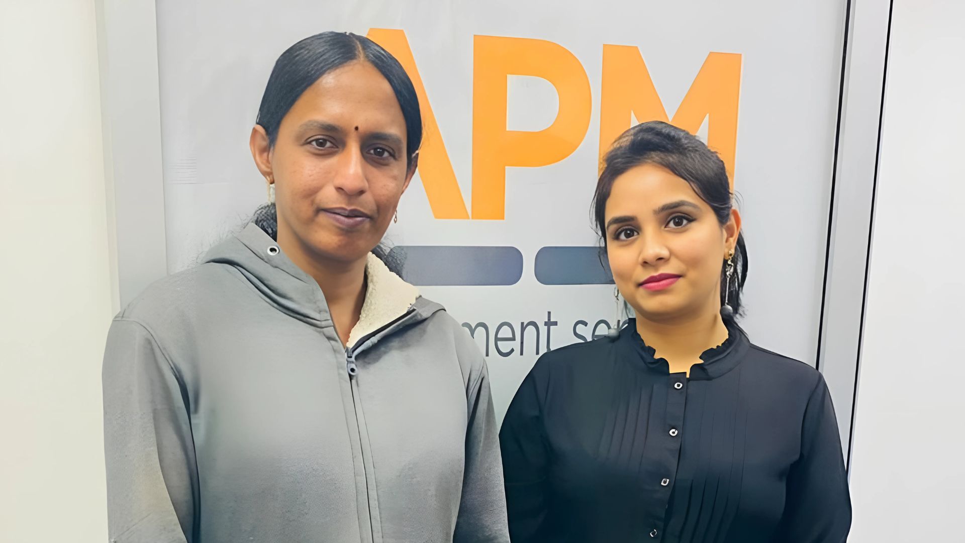 Anita and Harpreet smile together next to an APM sign