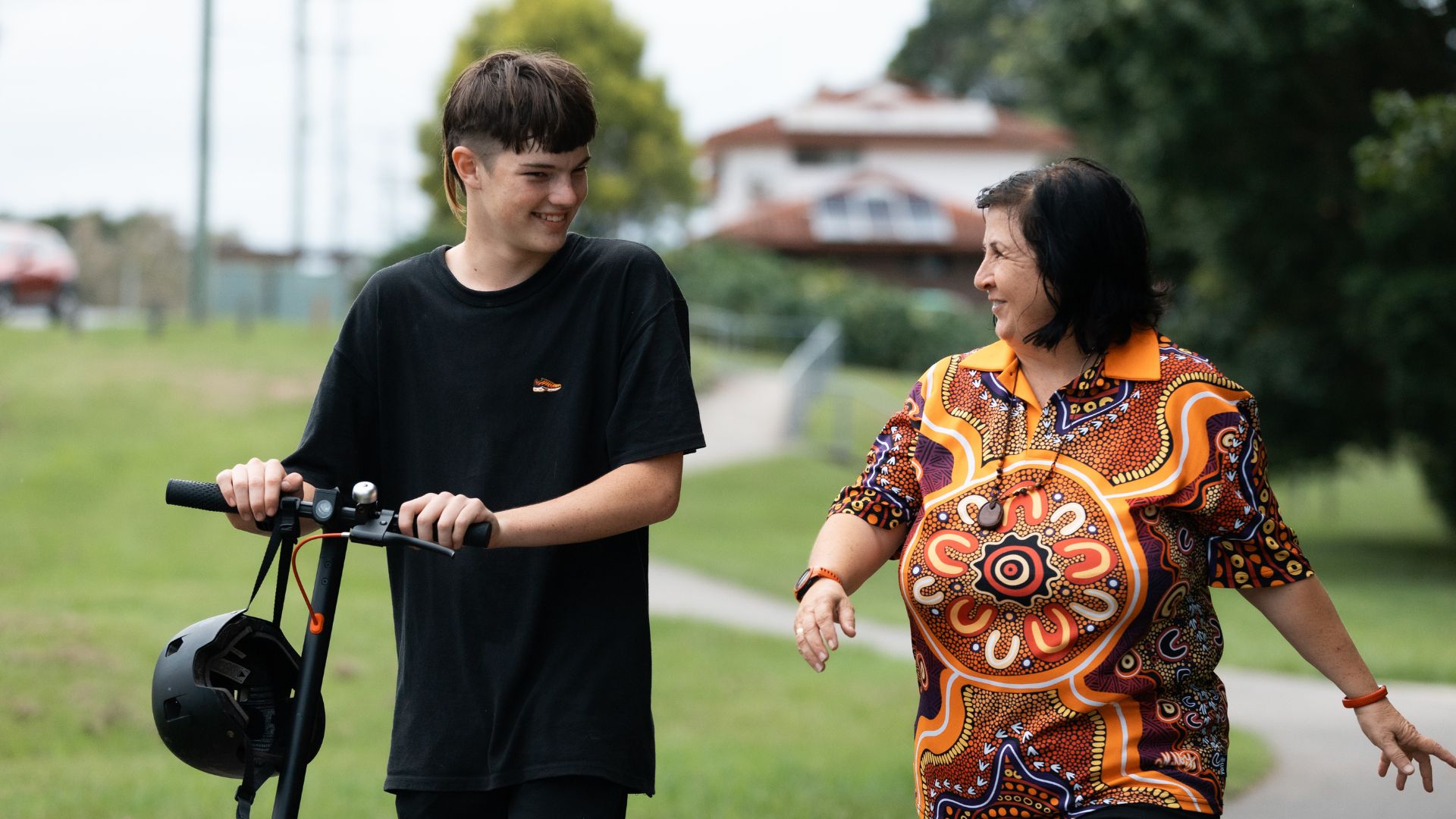 William is walking outdoors while holding an electric scooter, accompanied by a support worker from Everyday Independence who is smiling and talking to him.