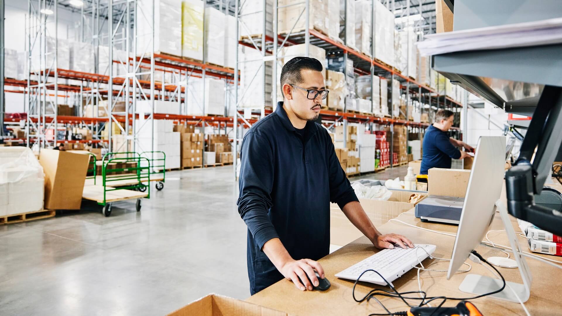 A man standing at a desk working on a computer in a warehouse