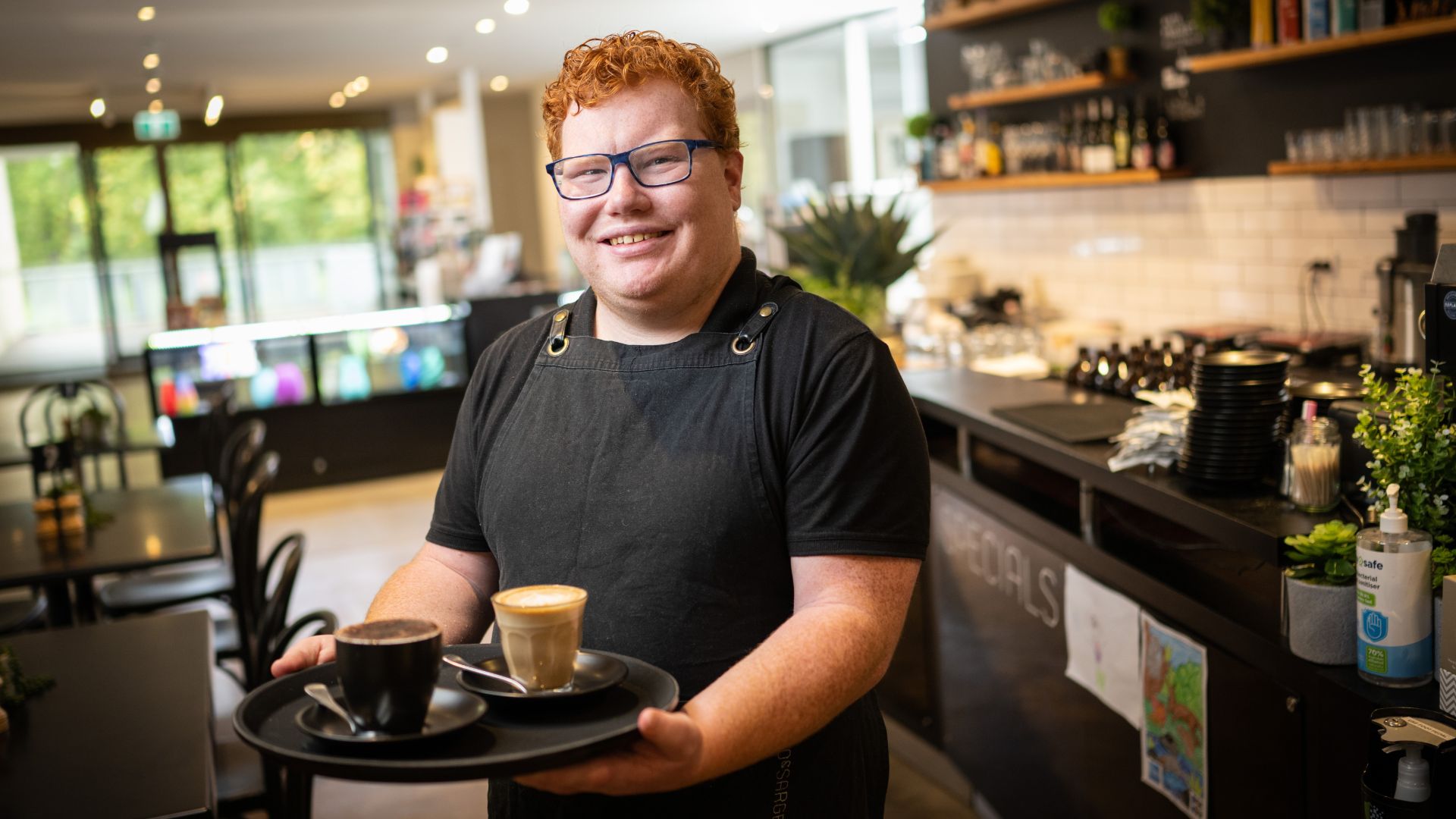 Cafe worker holding a tray with two cups of coffee at a service counter, highlighting the benefits of hiring people with disability in customer service roles