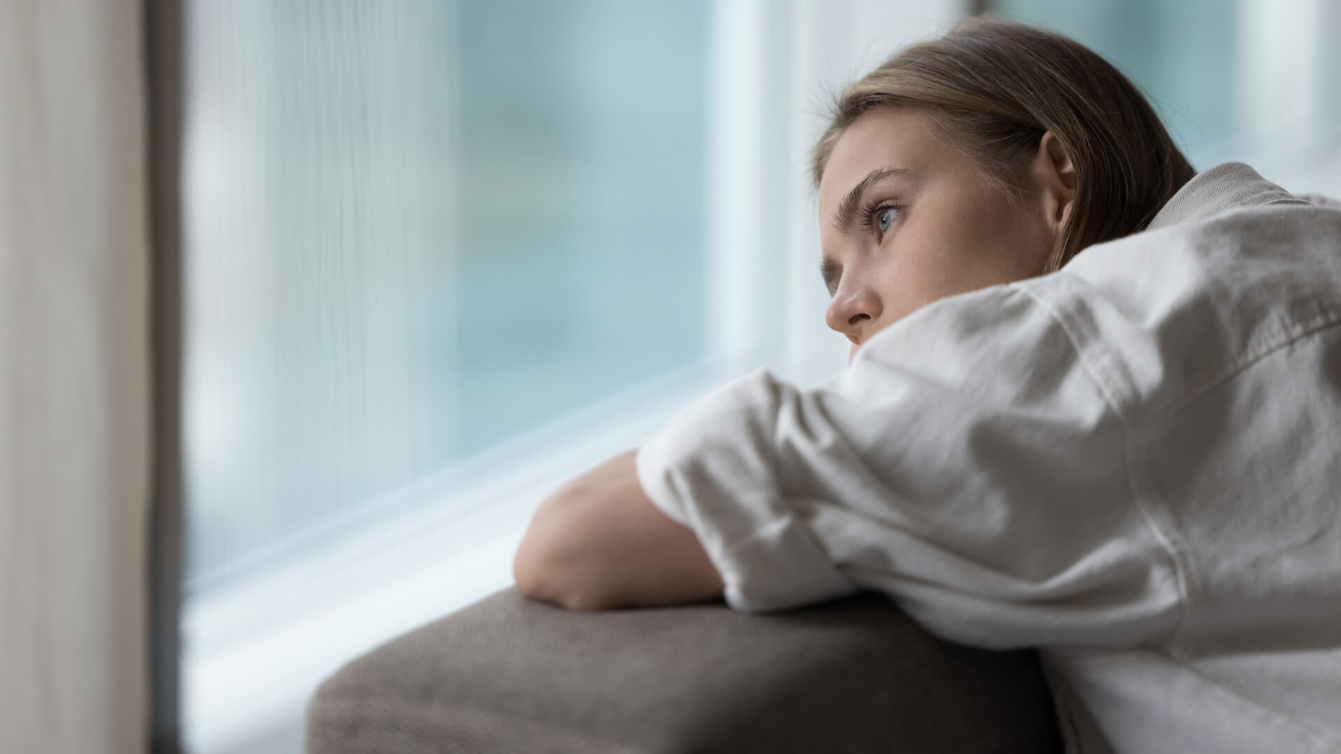 Young woman leaning on a couch and gazing out a window, looking thoughtful or concerned