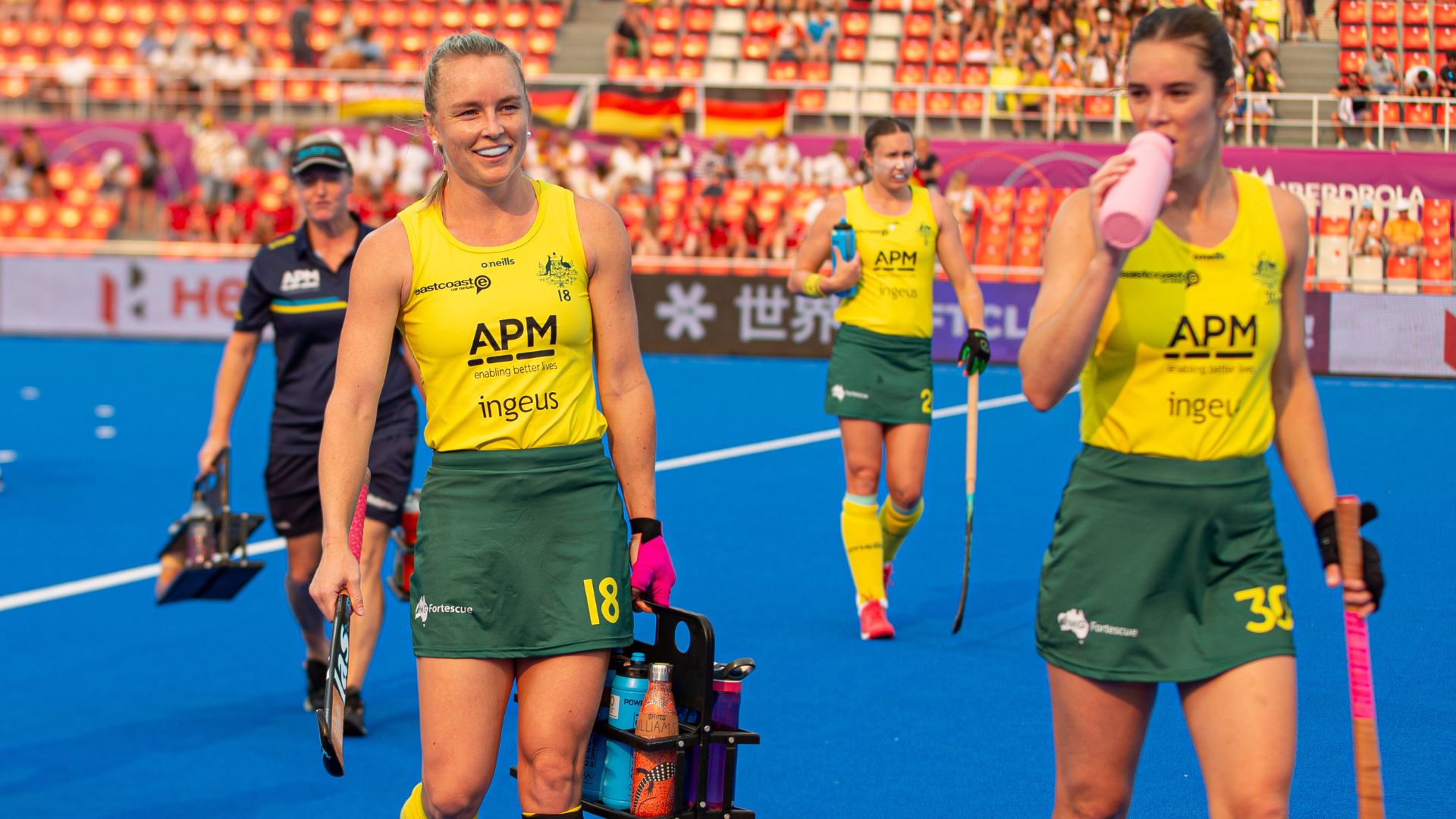 Australian women’s hockey team players in yellow and green uniforms walking on a blue turf after a match, carrying sticks and water bottles. APM and Ingeus logos are visible on the uniforms. Stadium seating and banners in the background.