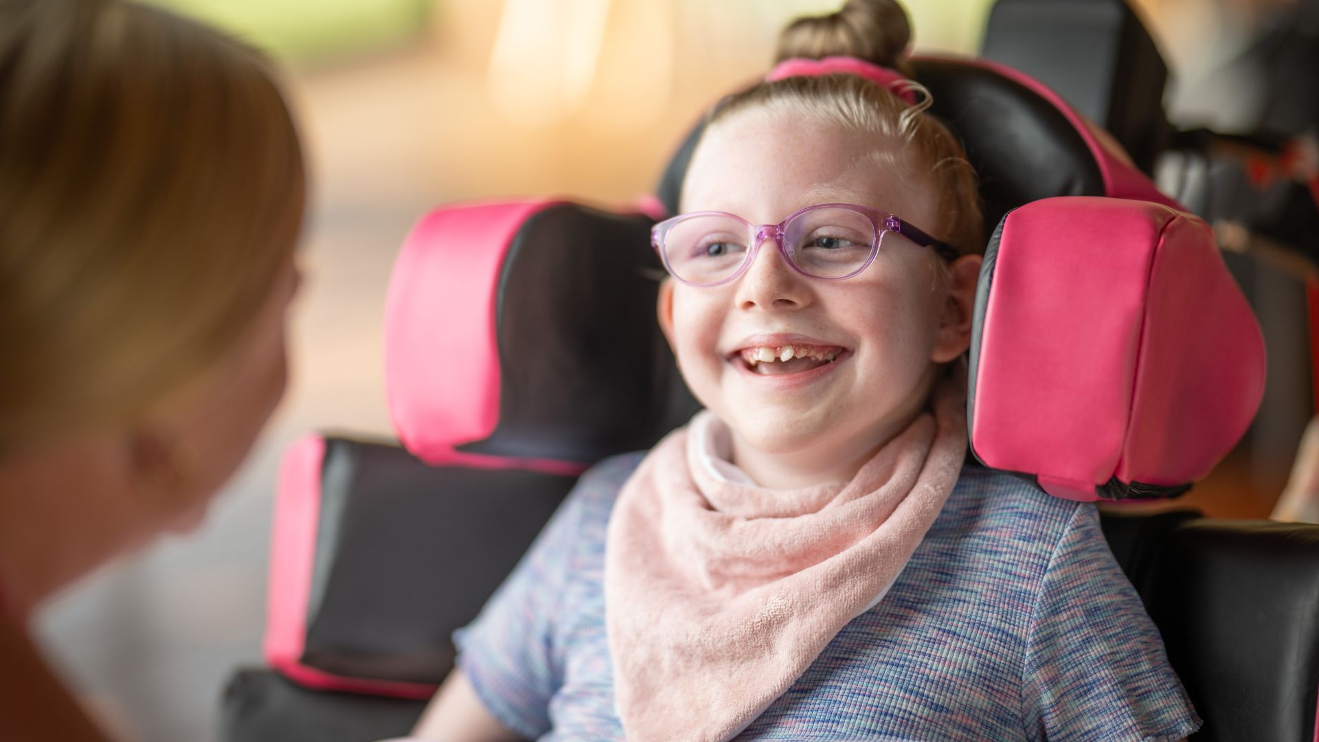 Emily is seated in a specialized wheelchair with pink head supports and a soft pink bib, facing an adult who is partially visible. The image focuses on Emily’s assistive equipment, emphasizing care for children with high support needs.