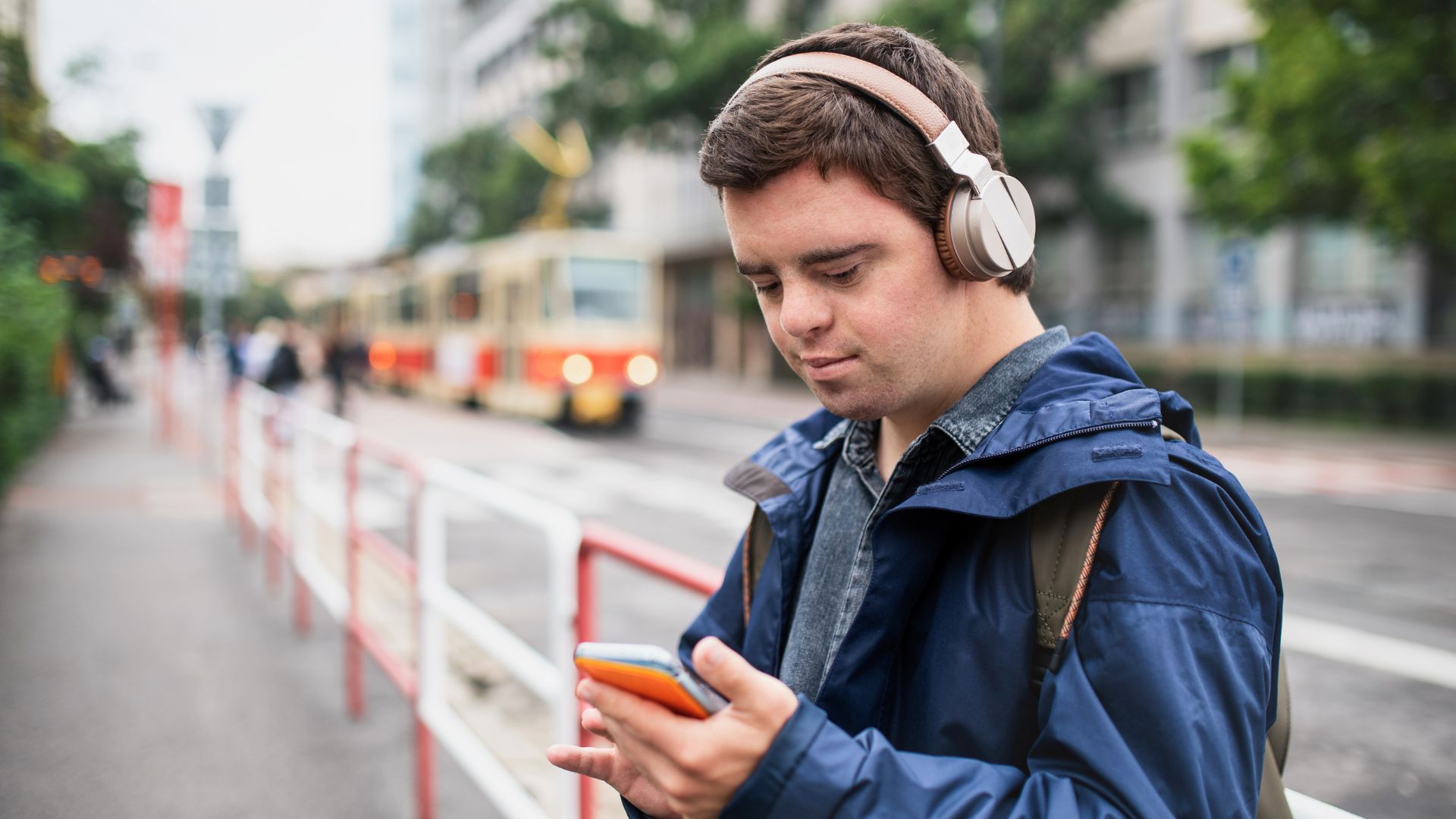 Man wearing headphones looking at his smartphone while standing near a tram stop on a city street.