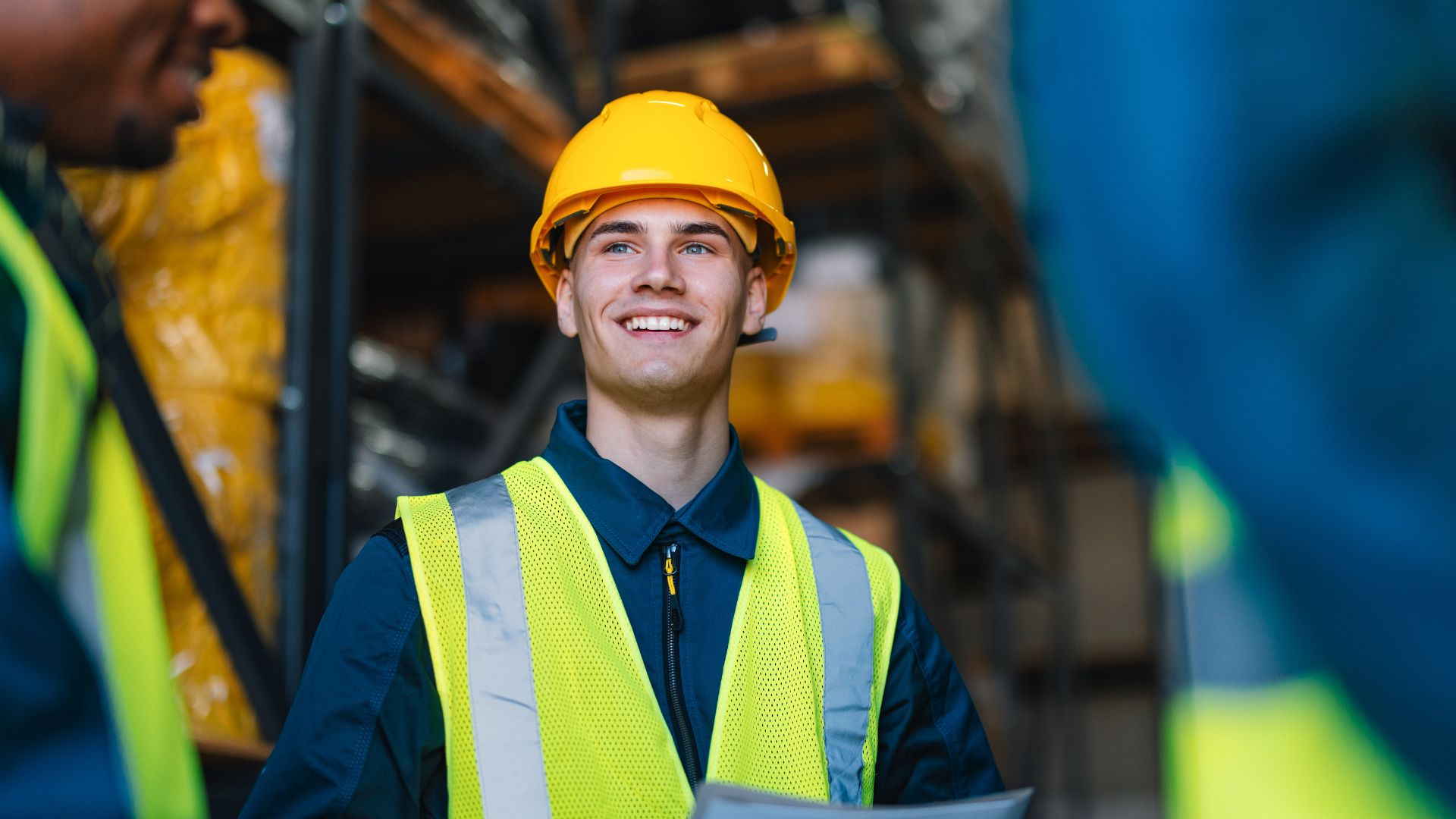 A worker wearing a hard hat and high-visibility vest smiles while standing in a warehouse, with shelves and equipment visible in the background.
