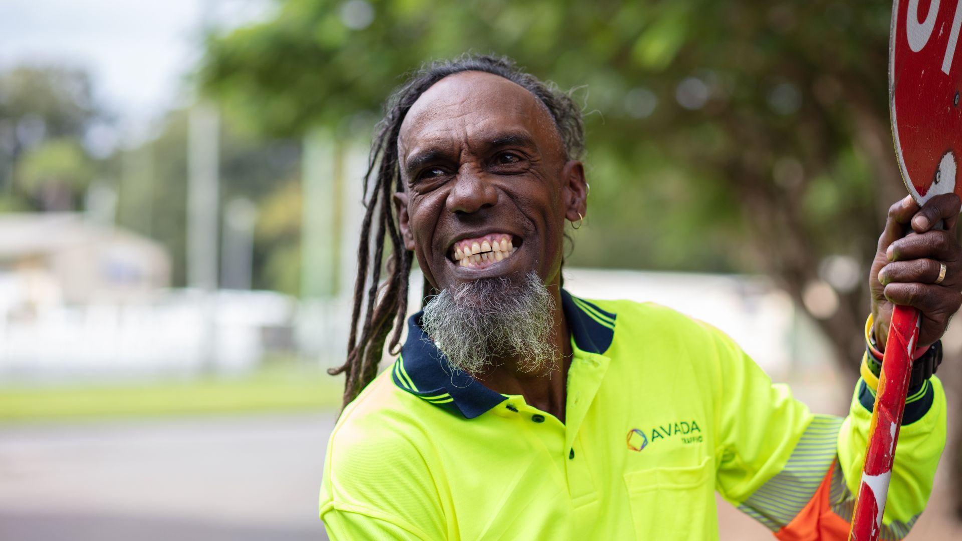 Close-up of Workforce Australia participant Adam holding a stop sign and smiling at the camera while wearing high-visibility traffic control attire outdoors.