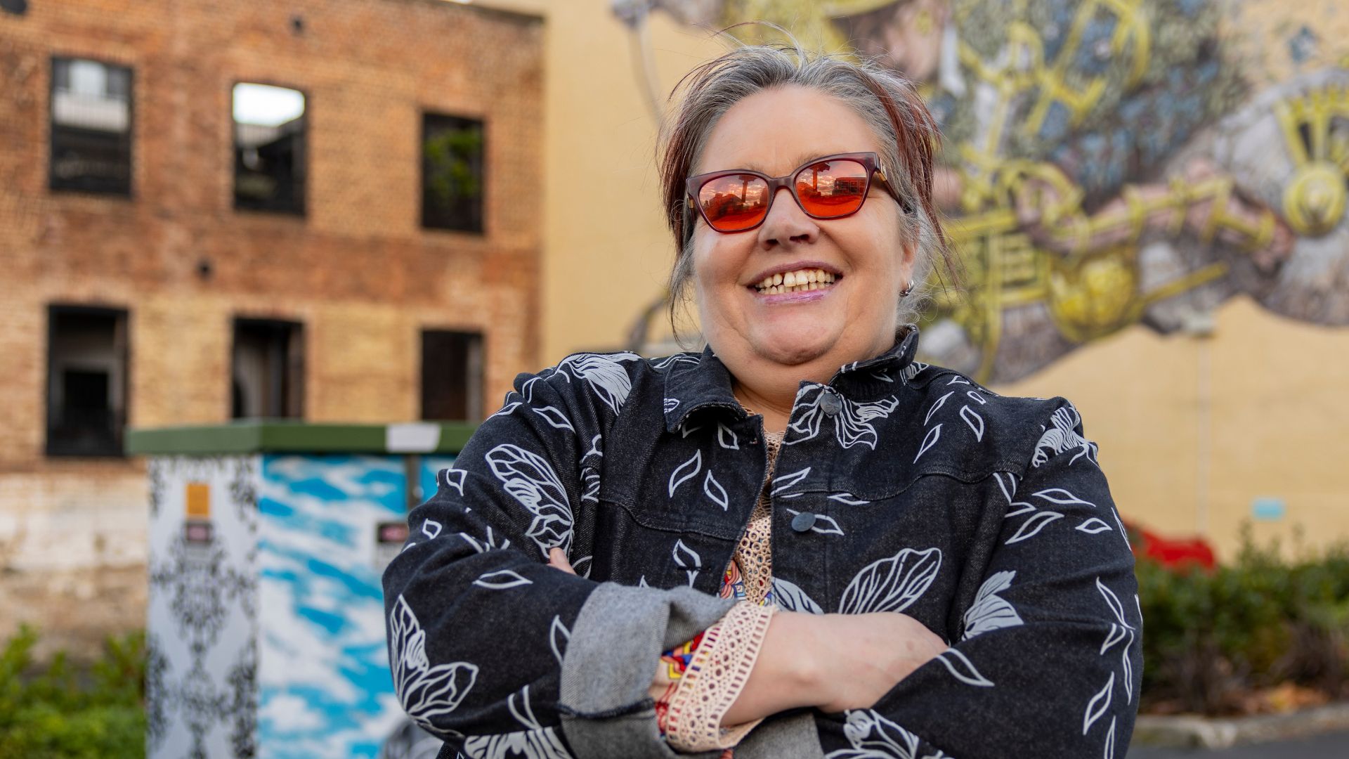 A woman stands smiling outside infront of a wall mural