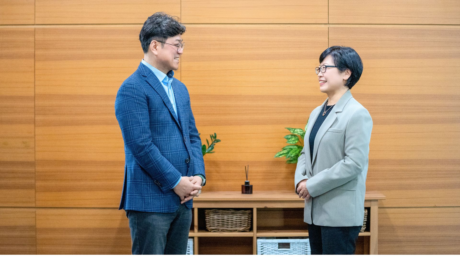 Sungjun Kim standing and speaking with career consultant Meeae Kim in a modern office setting with wooden panel walls and indoor plants, representing career transition support.