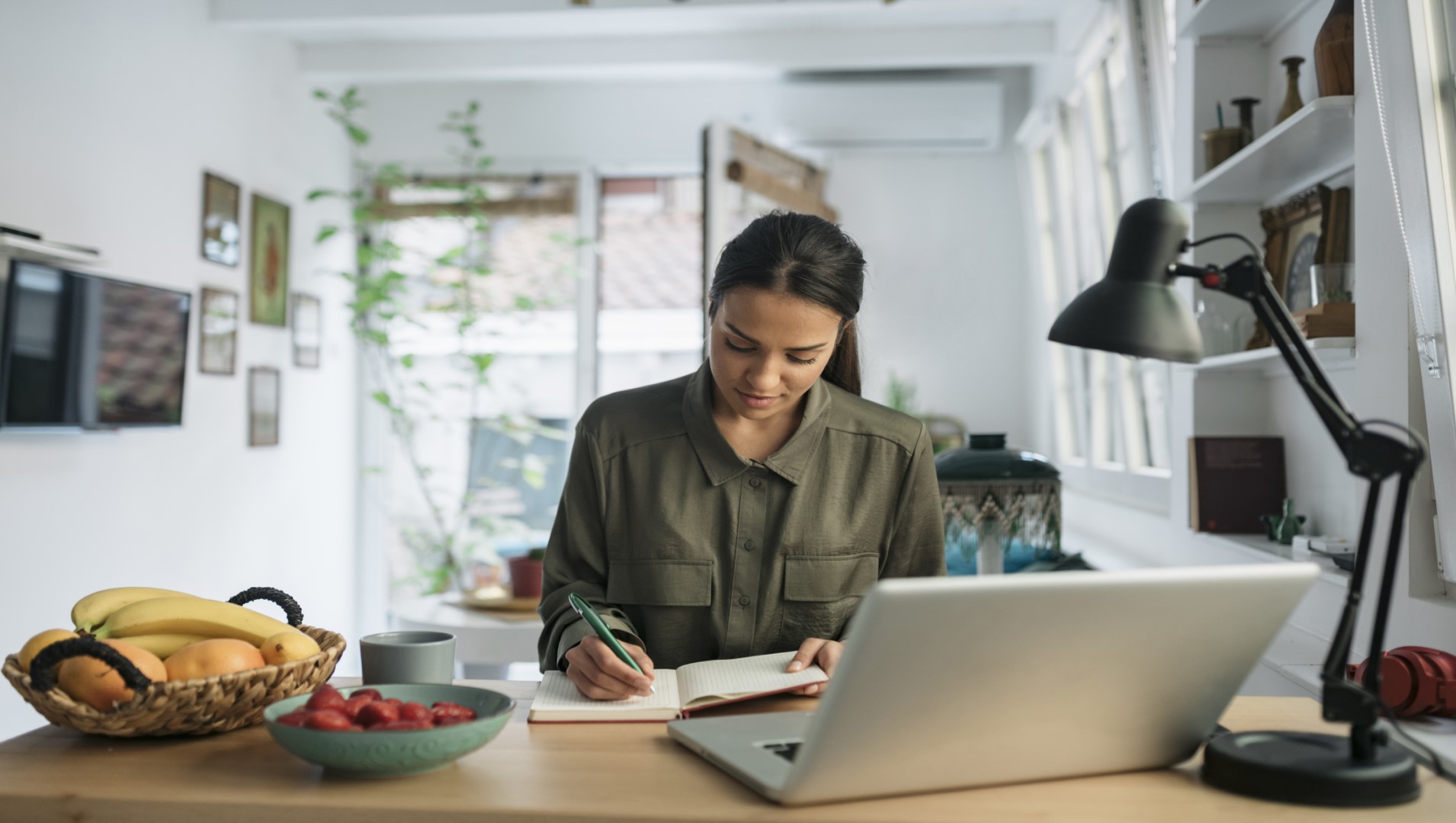 A woman sitting at a desk in a home office writing in a notebook